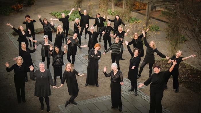 En grupp kvinnor i svarta kläder står utspridda och gör glada, sprudlande gester, lite som dans med viftande händer. I mitten står en kvinna med rött hår, stilla, hon är blickfånget.