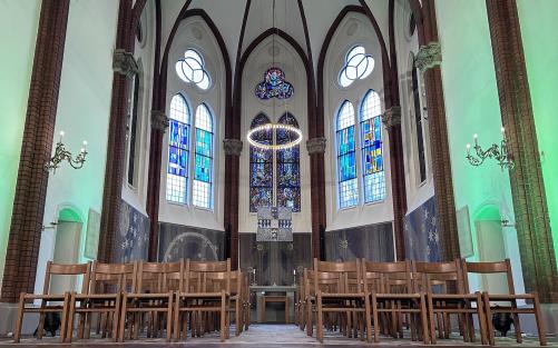 Längst fram i koret i en kyrka från 1800-talet. Vi ser målade glasfönster, både gammeldags och moderna, stolar, pelare i röd tegelsten, vita, putsade väggar upplysta i grönt, ett altare och ett massivt kvadratiskt glaskors.