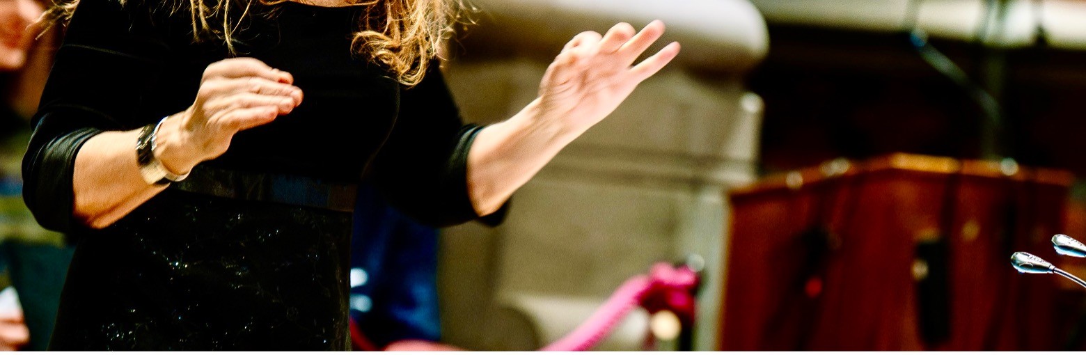 Du ser händerna på någon som dirigerar. Personen har en svart tröja och blå byxor.