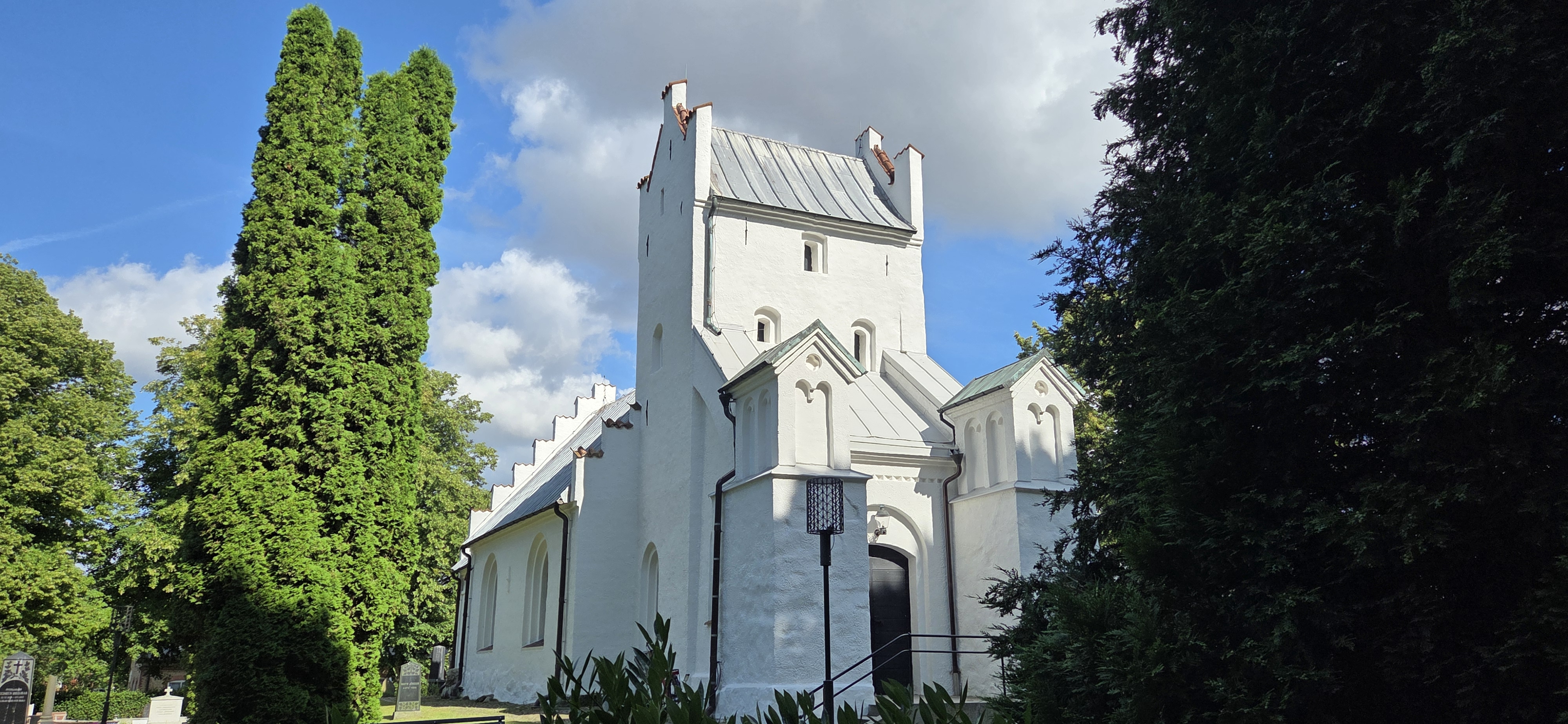 Vit kyrka mot en blå himmel. Omgiven av grönskande buskar och träd