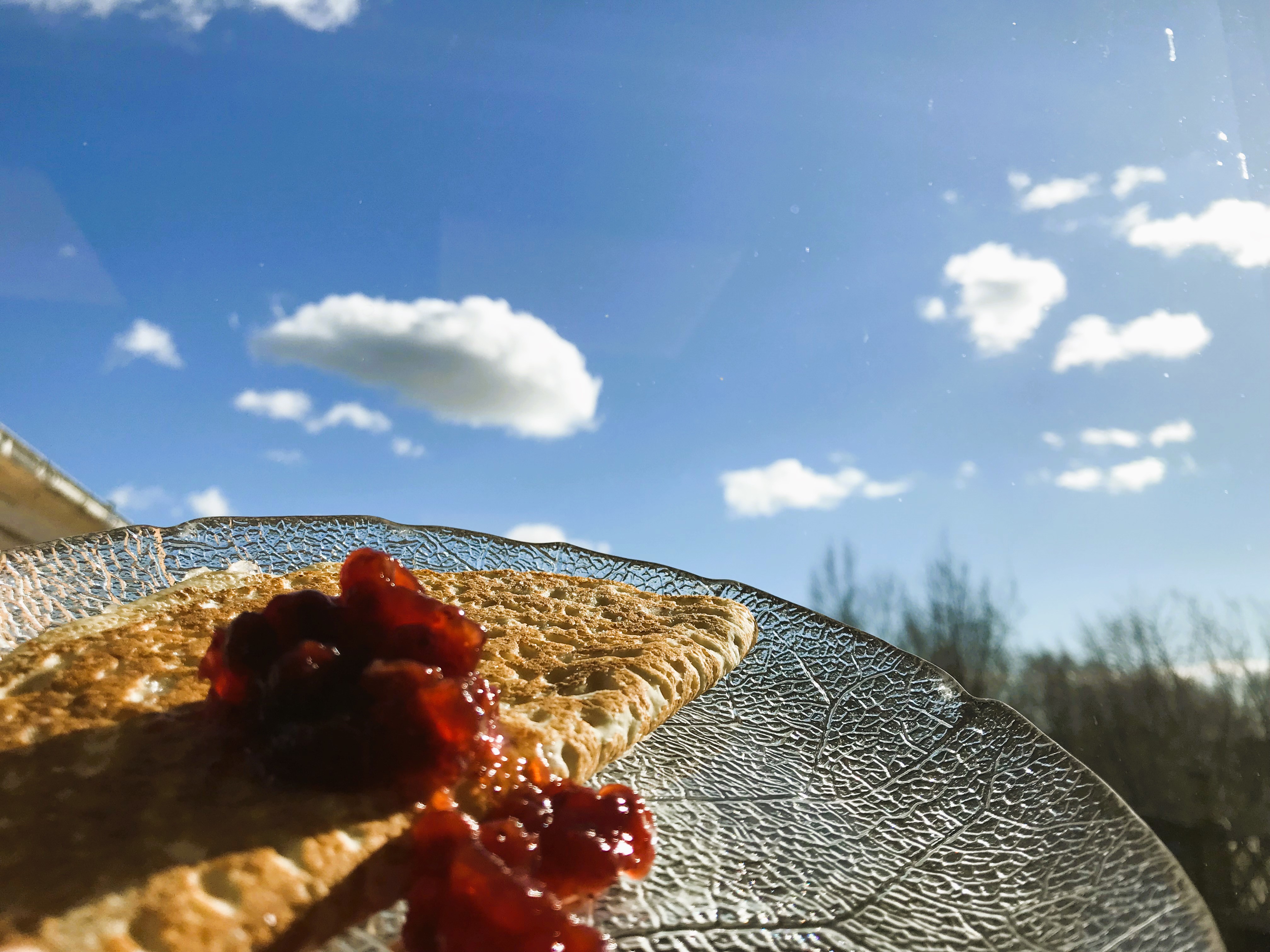 Pannkaka med sylt på en glastallrik hålls upp i solljus mot en blå, moln beströdd himmel
