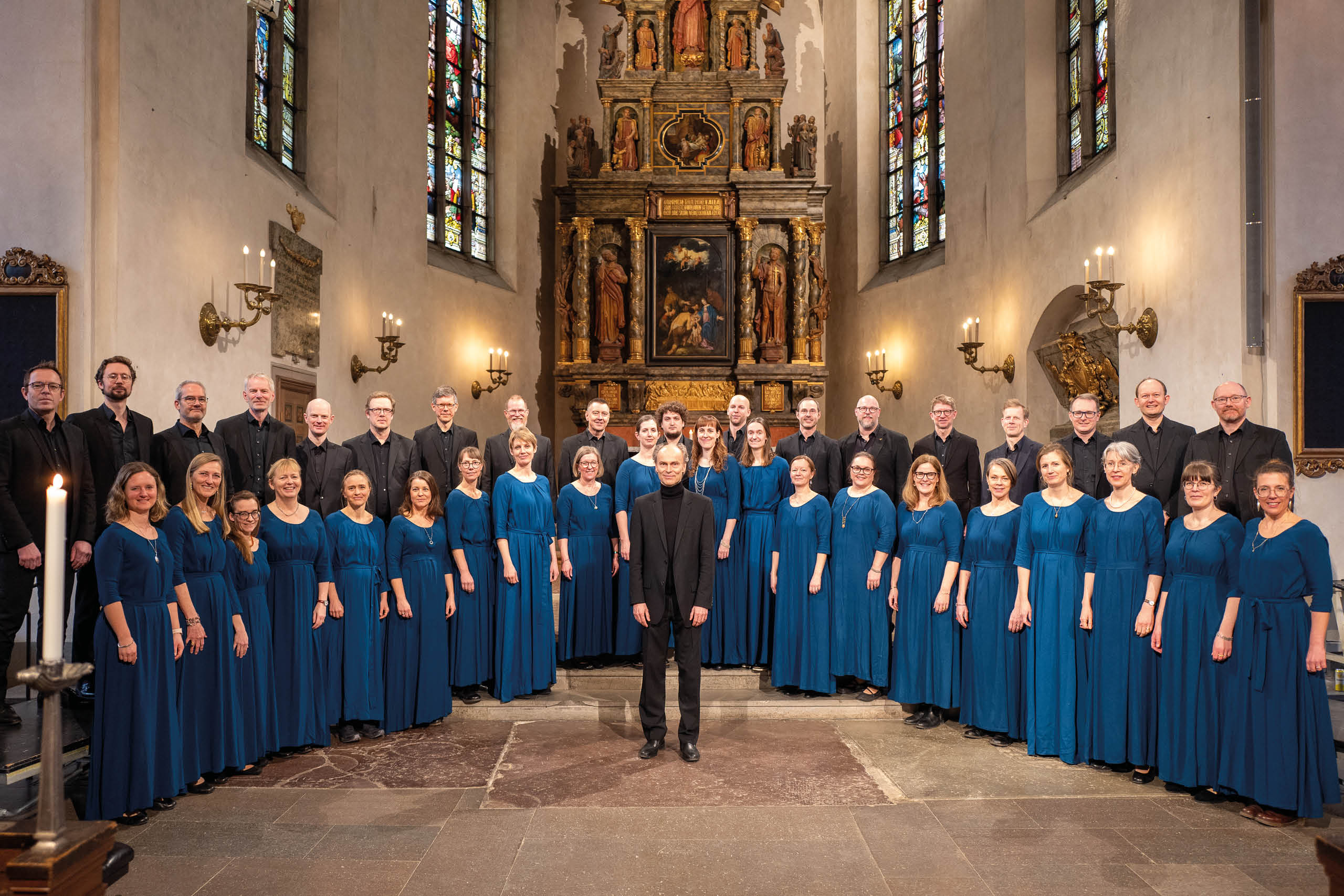 Kören står uppställda i en kyrka framför altaret. Alla kvinnor har blåa klänningar och männen svarta kostymer.