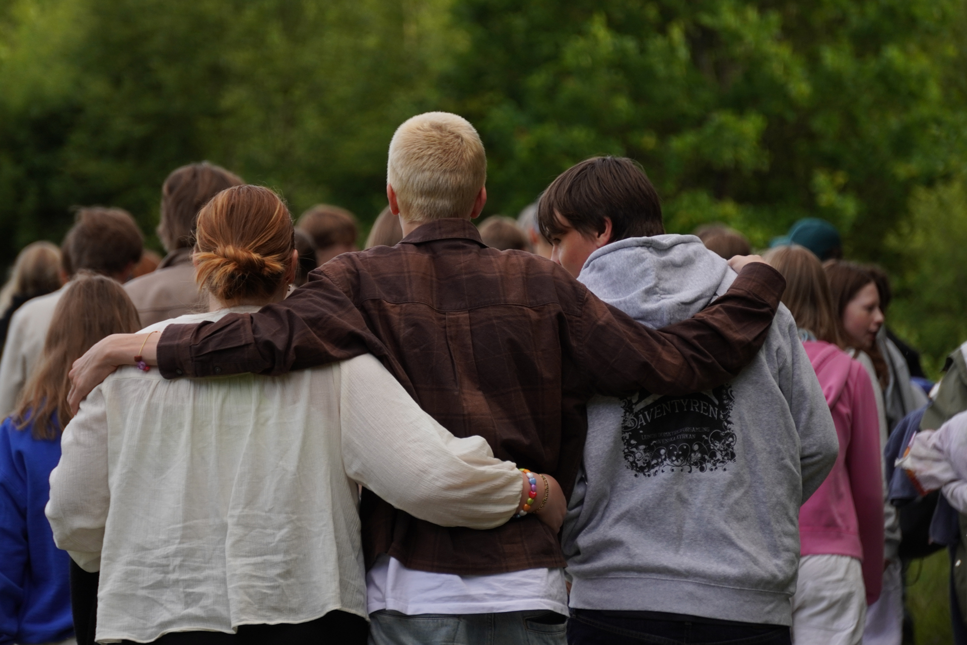 En grupp ungdomar går utomhus mot en träddunge. En person håller sina armar om axlarna på två andra. 
