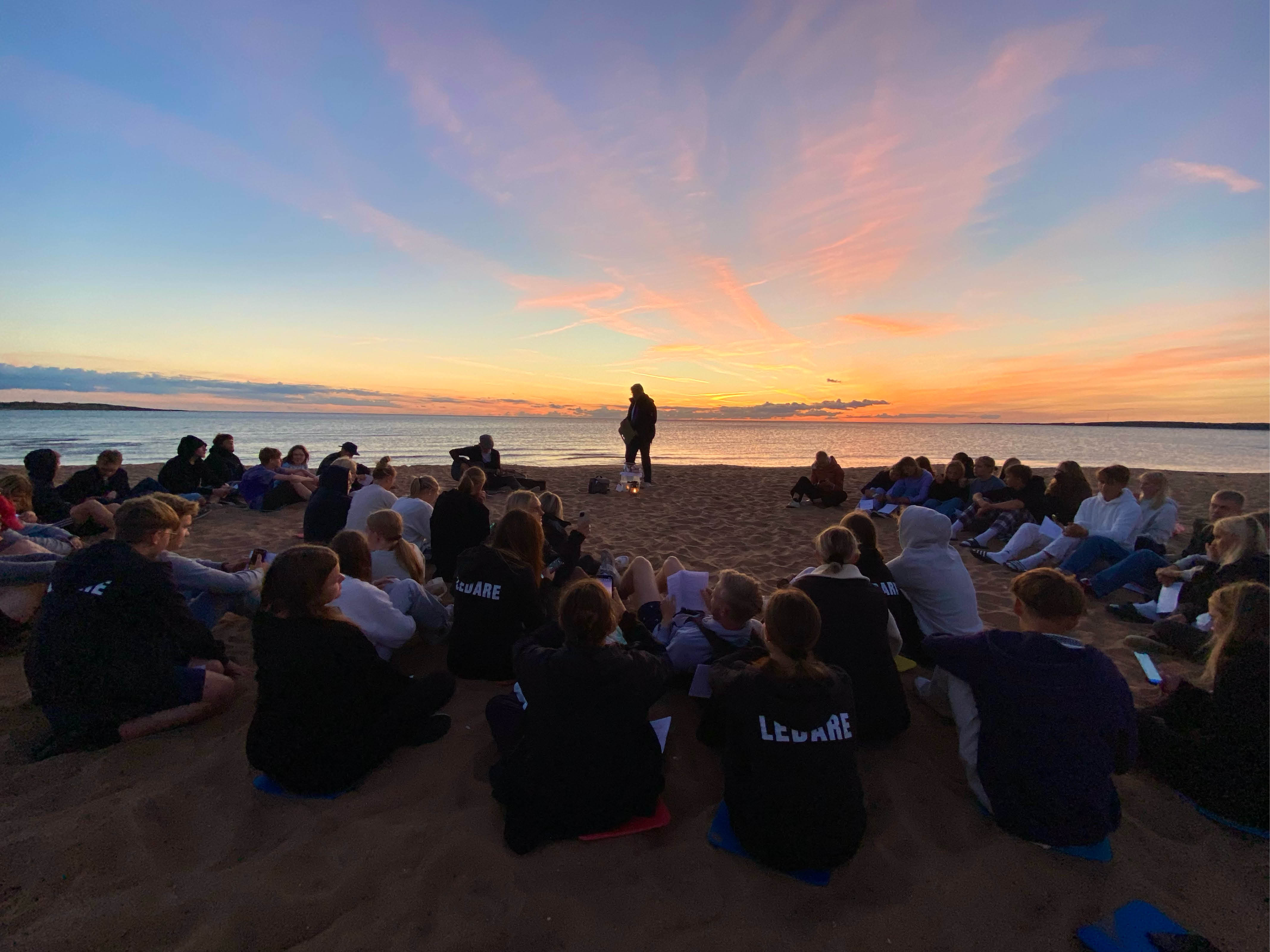 En grupp konfirmander och ledare sitter i en stor ring på stranden. Himlen färgas blå, orange och rosa av solnedgången.