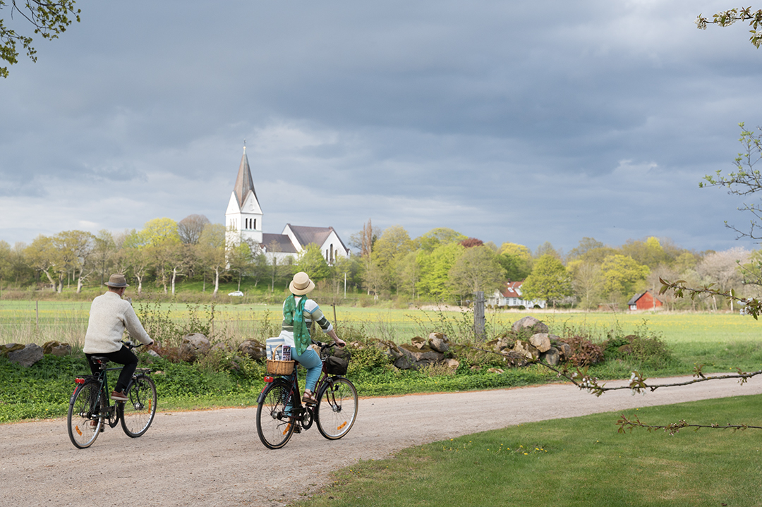 Två personer cyklar på en grusväg, i fjärran skymtar en vit kyrka.