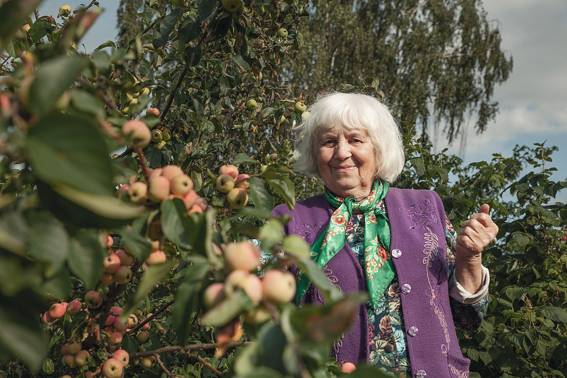 En glad äldre kvinna står bredvid ett dignande äppleträd i en trädgård.
