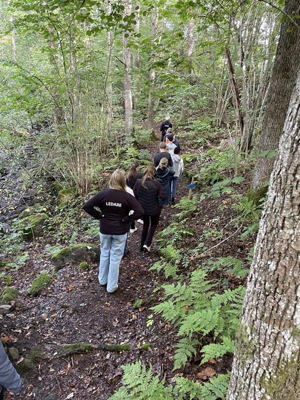 En rad med ungdomar går på en stig i skogen. Längst bak går en ungdom med en tröja där det står ledare. 
