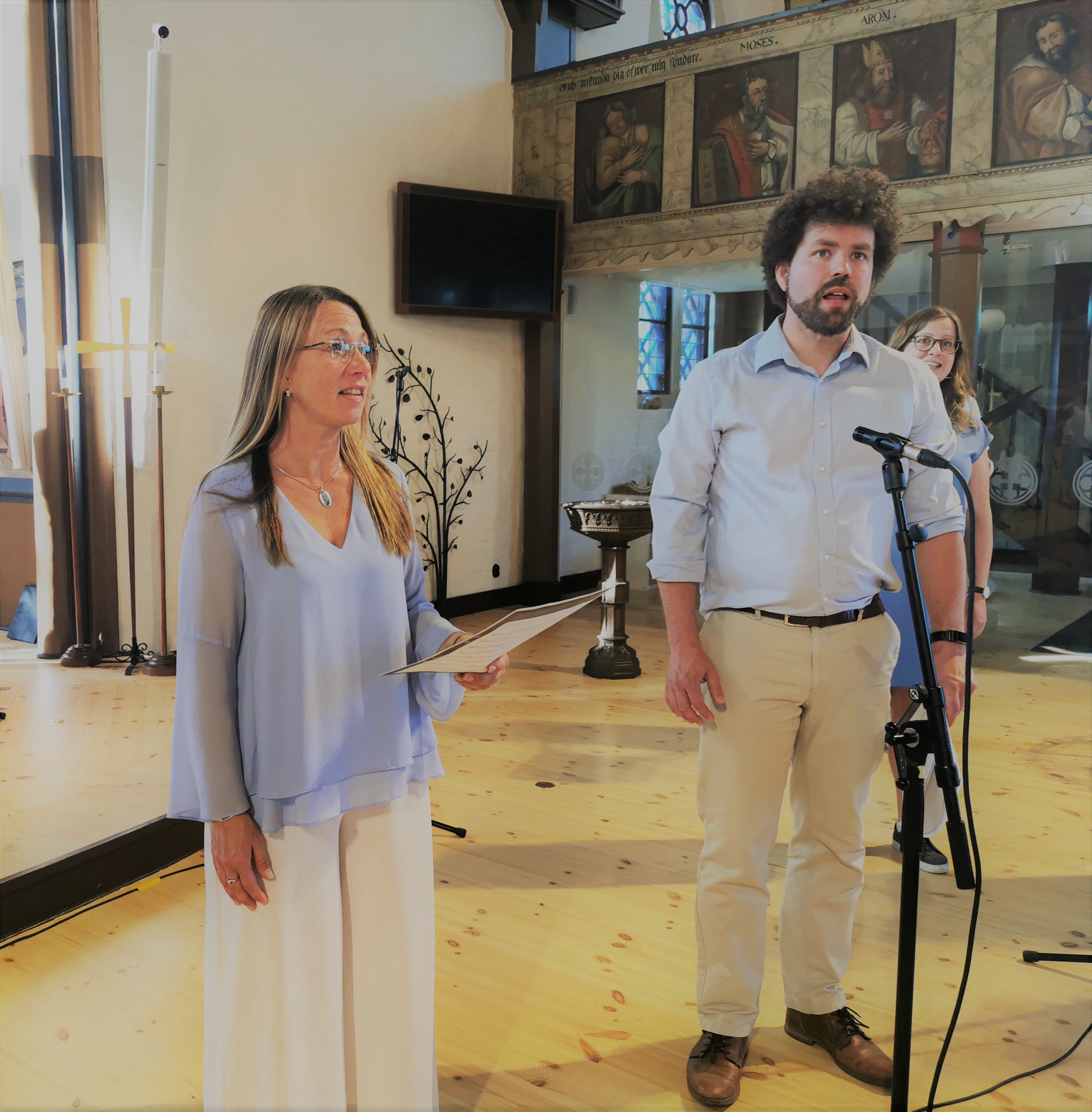 Två personer, en kvinna och en man, står och sjunger i Fässbergs kyrka. Bakom mannen skymtar ytterligare en kvinna som sjunger. 