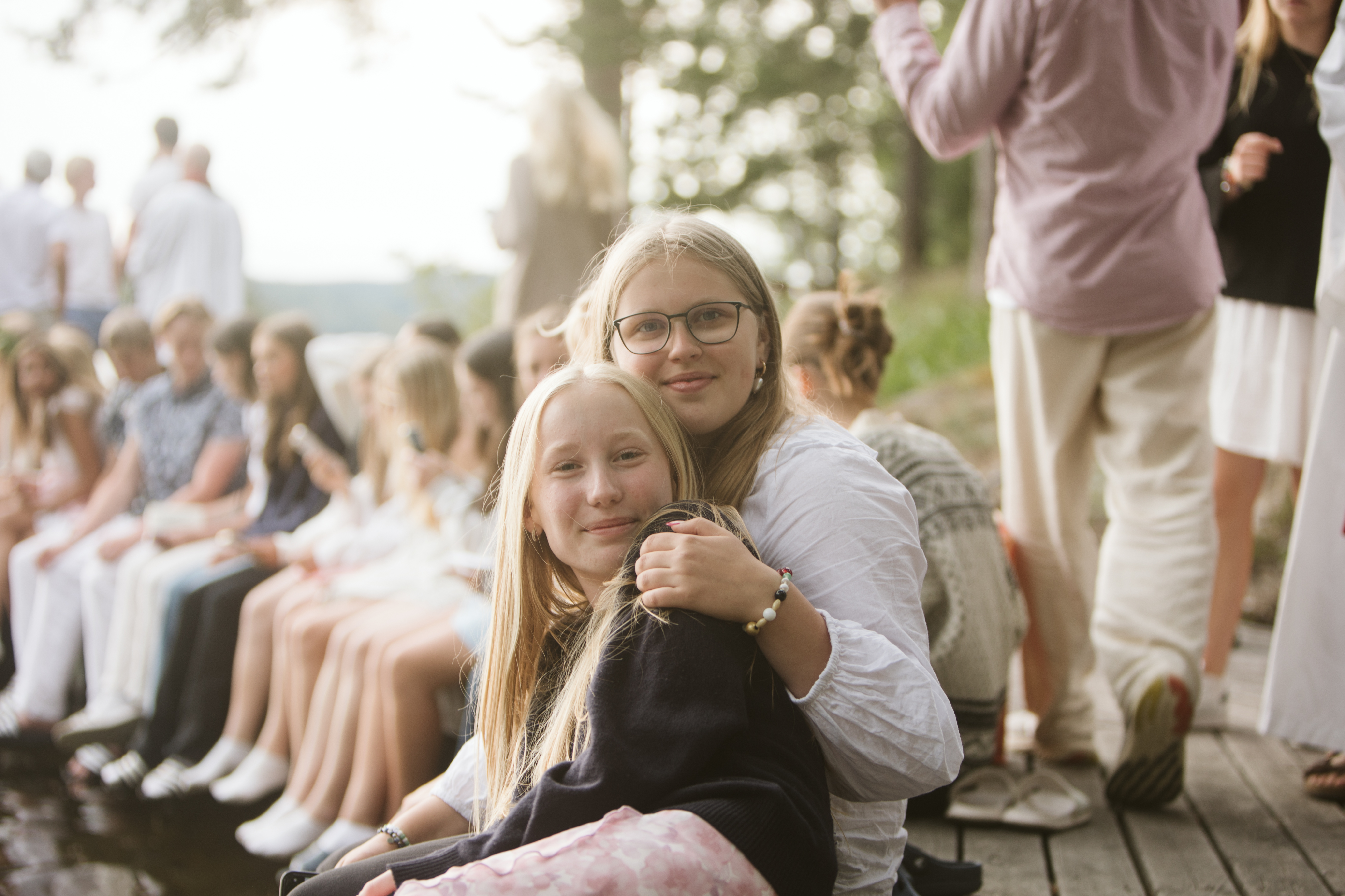 Två flickor, konfirmander, sitter tillsammans med andra ungdomar i bakgrunden. Båda tittar in i kameran. 