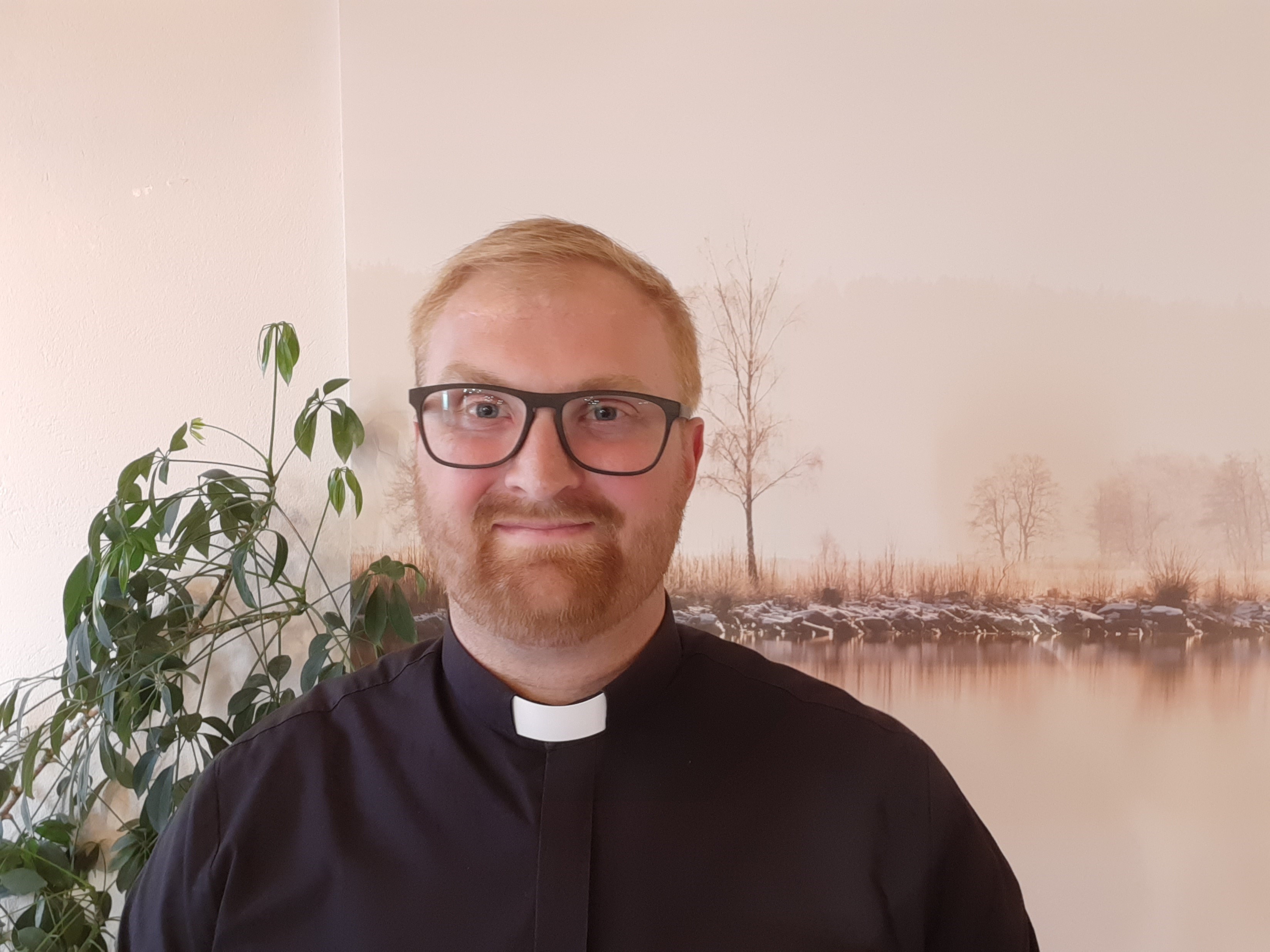 Alexander Dahlquist. Han står framför en vägg iförd en svart prästskjorta. Han har glasögon och skägg och ler mot kameran.