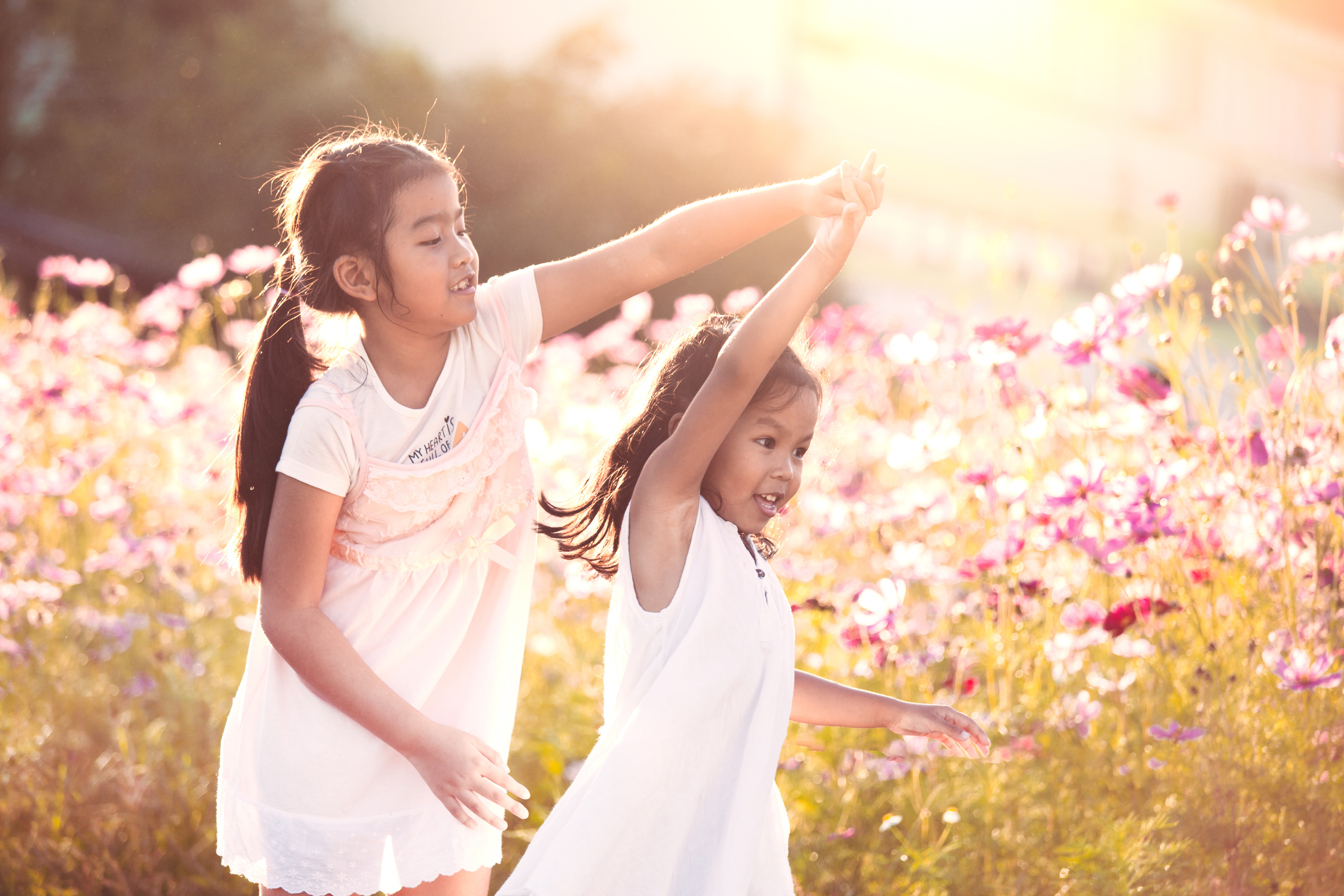 Två tjejer, ca 5 och 8 år, med mörkt hår och sommarkläder dansar eller leker på en sommaräng med rosa blommor.
