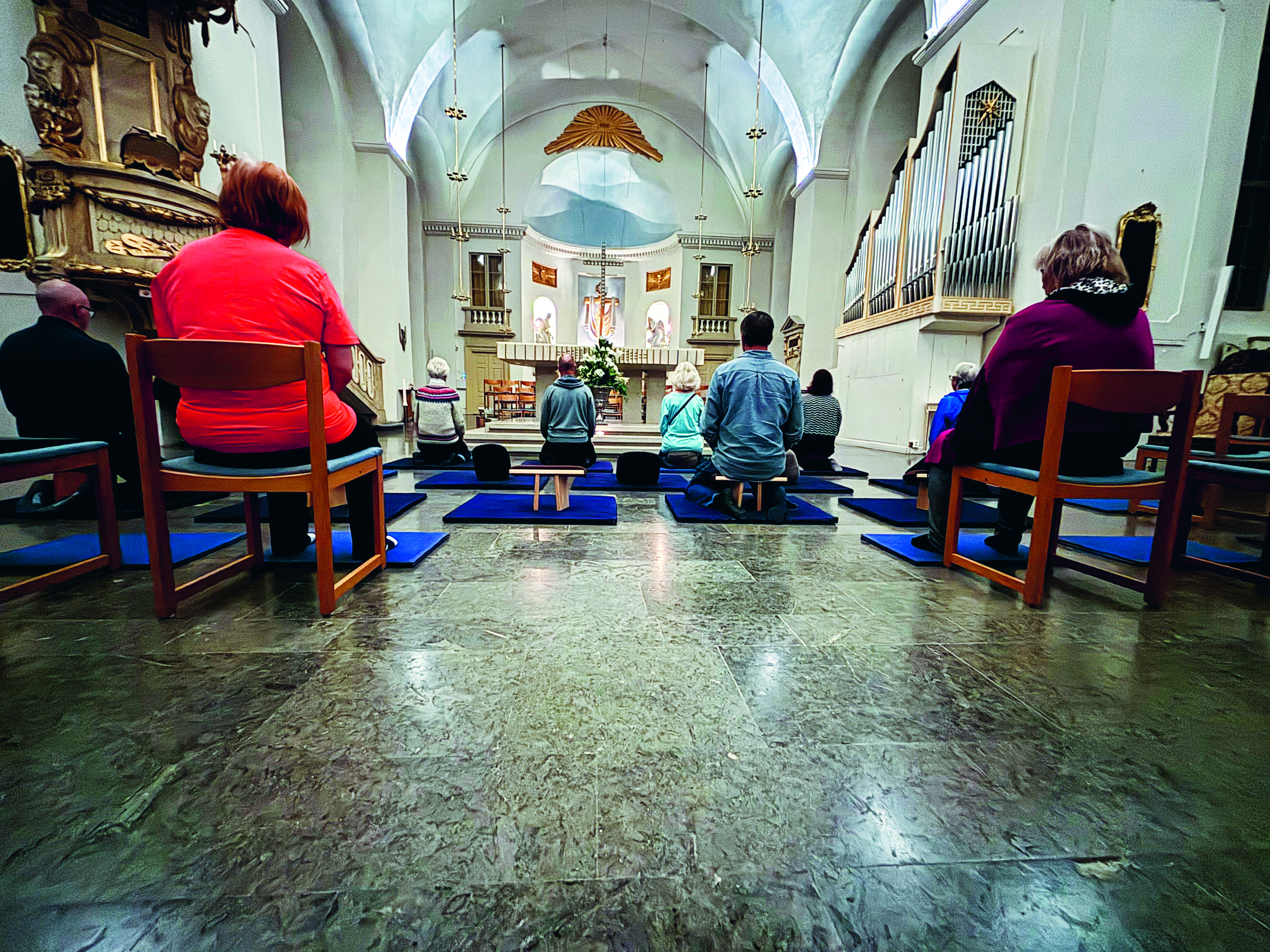 Man ser ett antal personer bakifrån, de sitter några på stol och några på kuddar på golvet i kyrkmiljö.