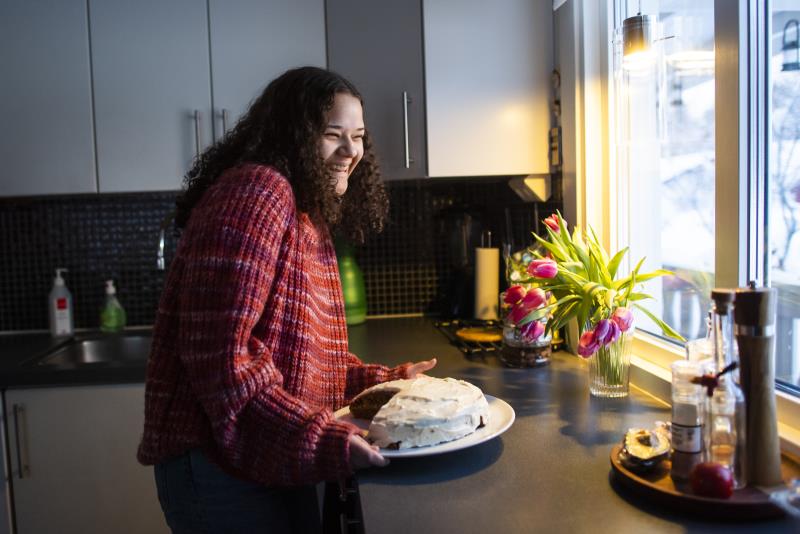 Ung kvinna håller i ett fat med en nybakt kaka. Hon står i köket framför fönstret där en vas med tulpaner är placerad. 