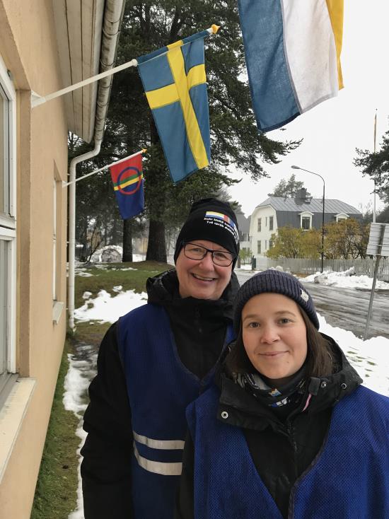 Under Tornedalens flagga står pedagogerna Rose-Marie Groth och Sara Pittja. De har den samiska och den svenska flaggan i bakgrunden. Sverigefinska flaggan syns inte men är vit med ett blått kors, en gul rand kantar korset.