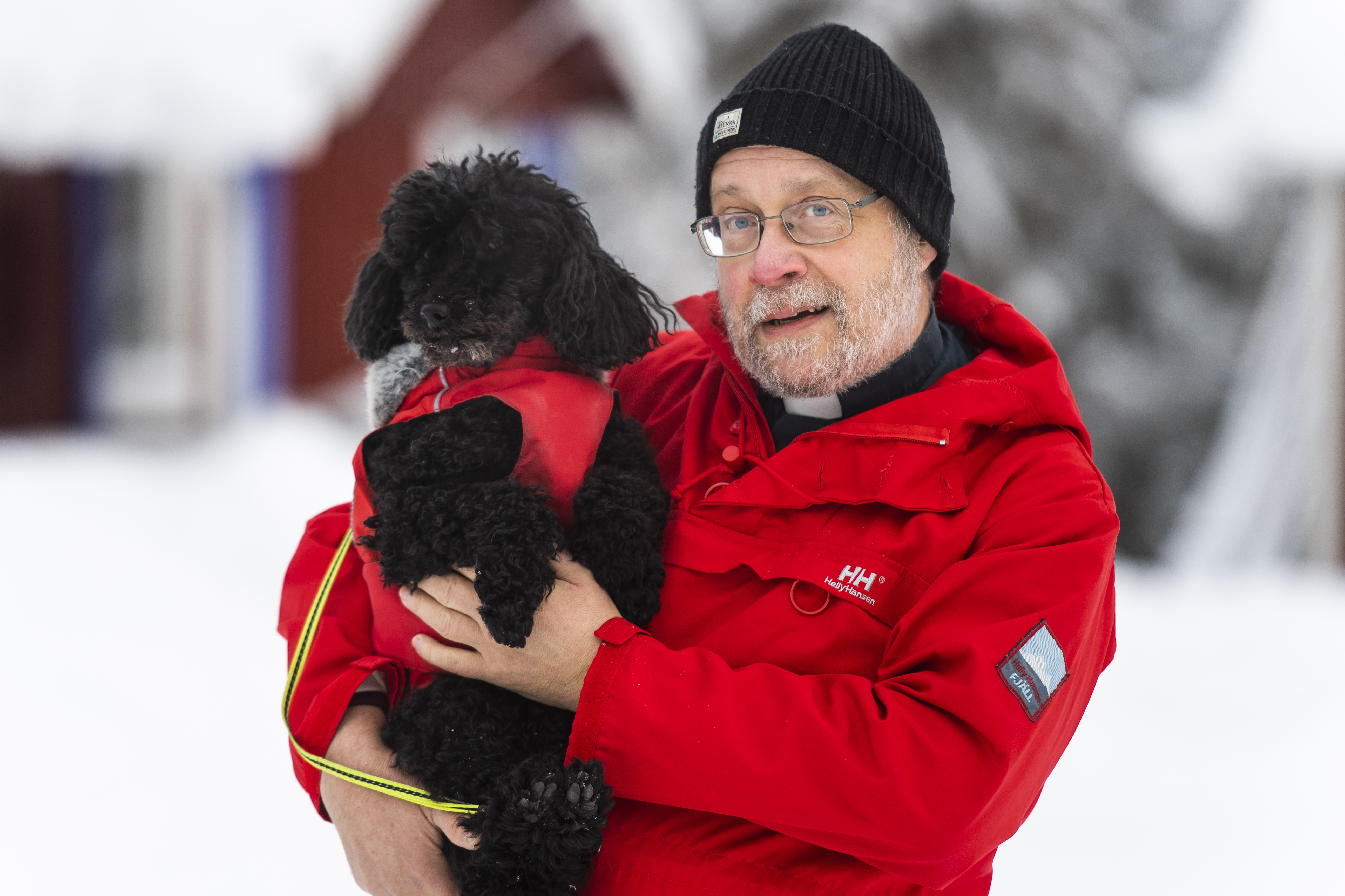 Man i röd anorak och svart mössa med en svart pudel i famnen. 