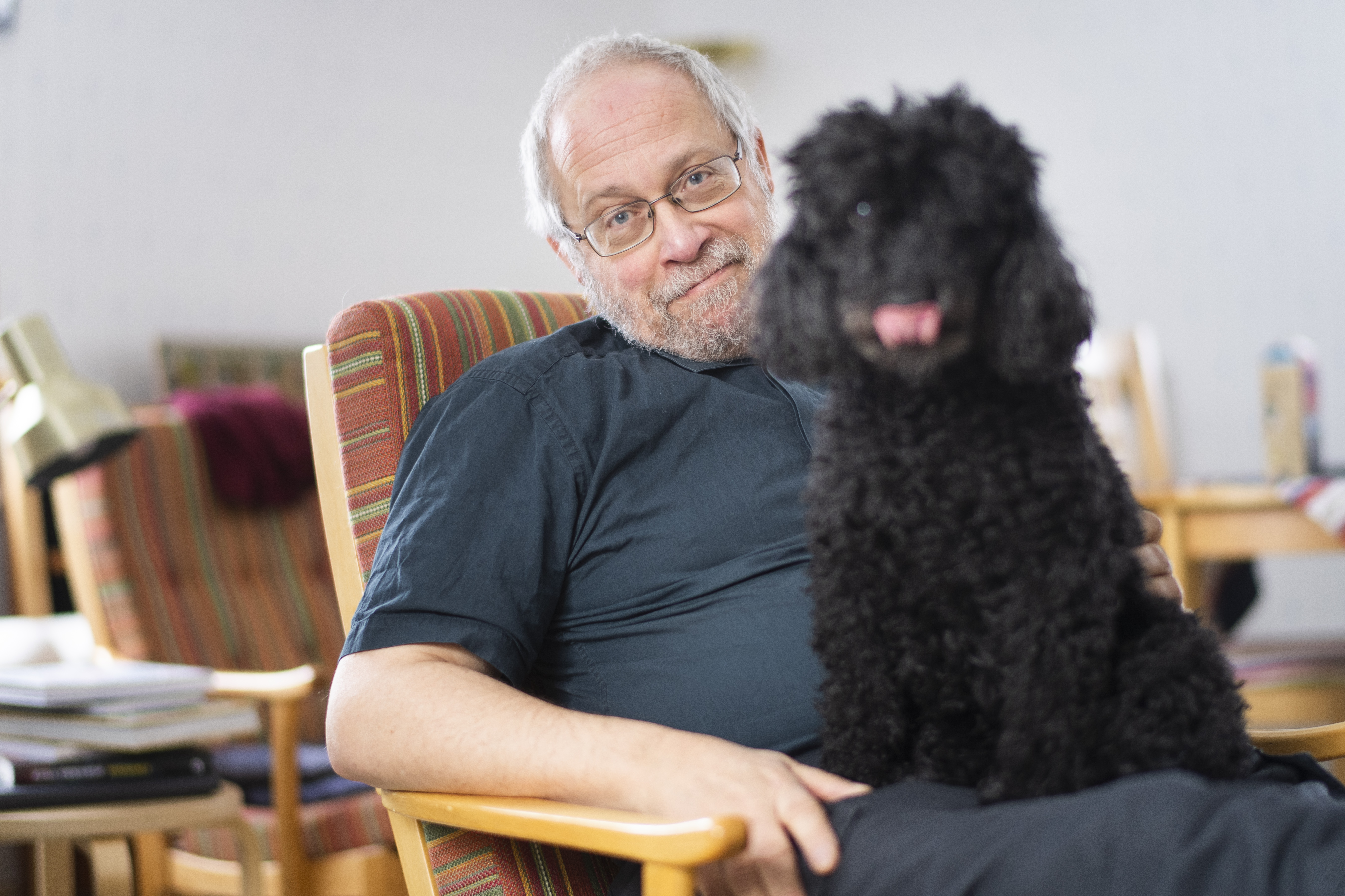 En glad man sitter i en fåtölj med sin svarta hund i famnen. 