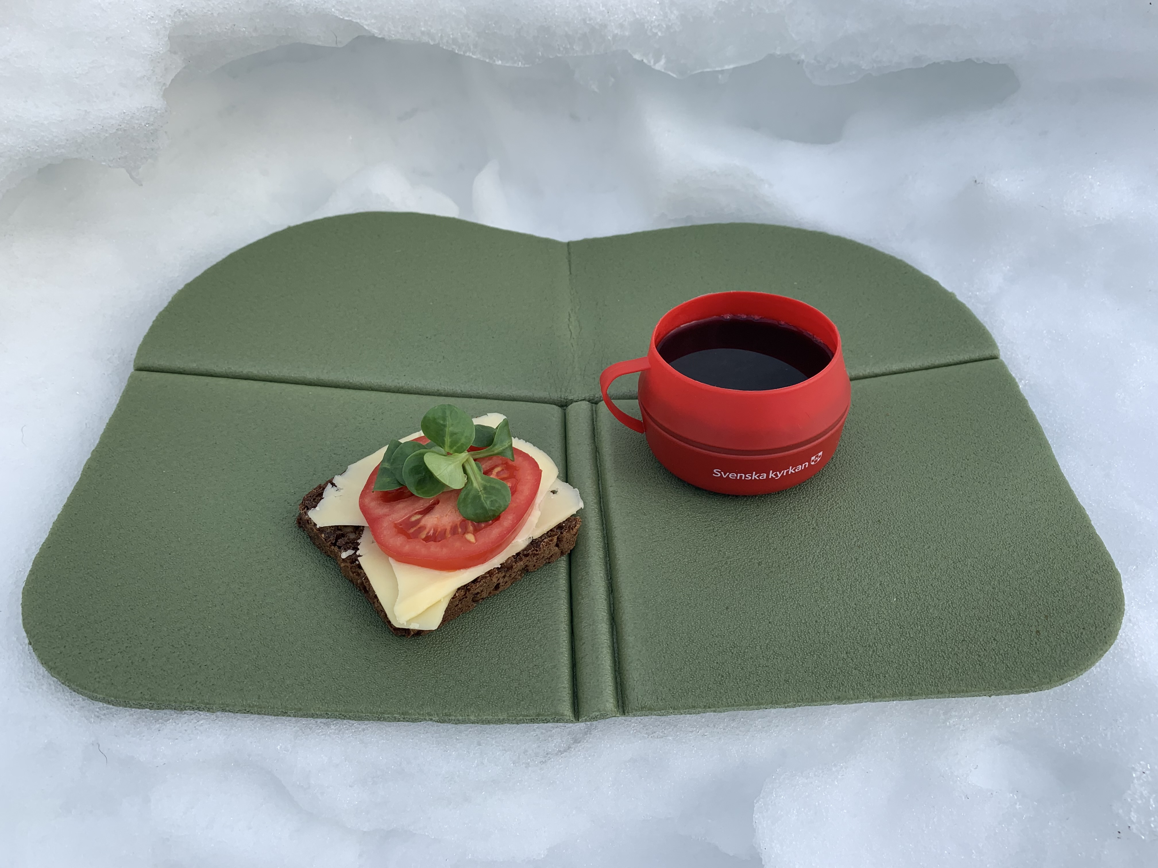 En mugg med kaffe och en smörgås på ett sittunderlag i snön..