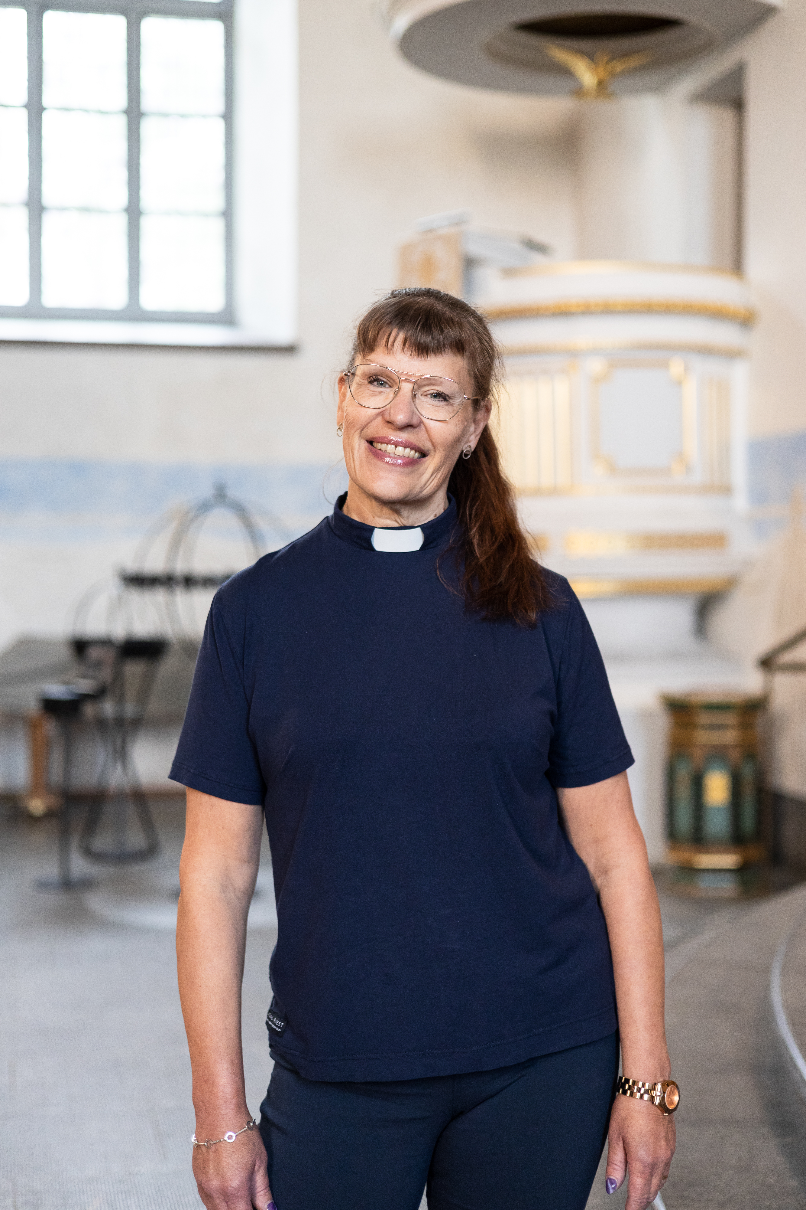 Person klädd i mörkblå prästskjorta står i en kyrkointeriör. I bakgrunden syns ett stort fönster, predikstol och kyrksalens inredning. Personen är placerad i bildens mitt och tittar mot kameran.