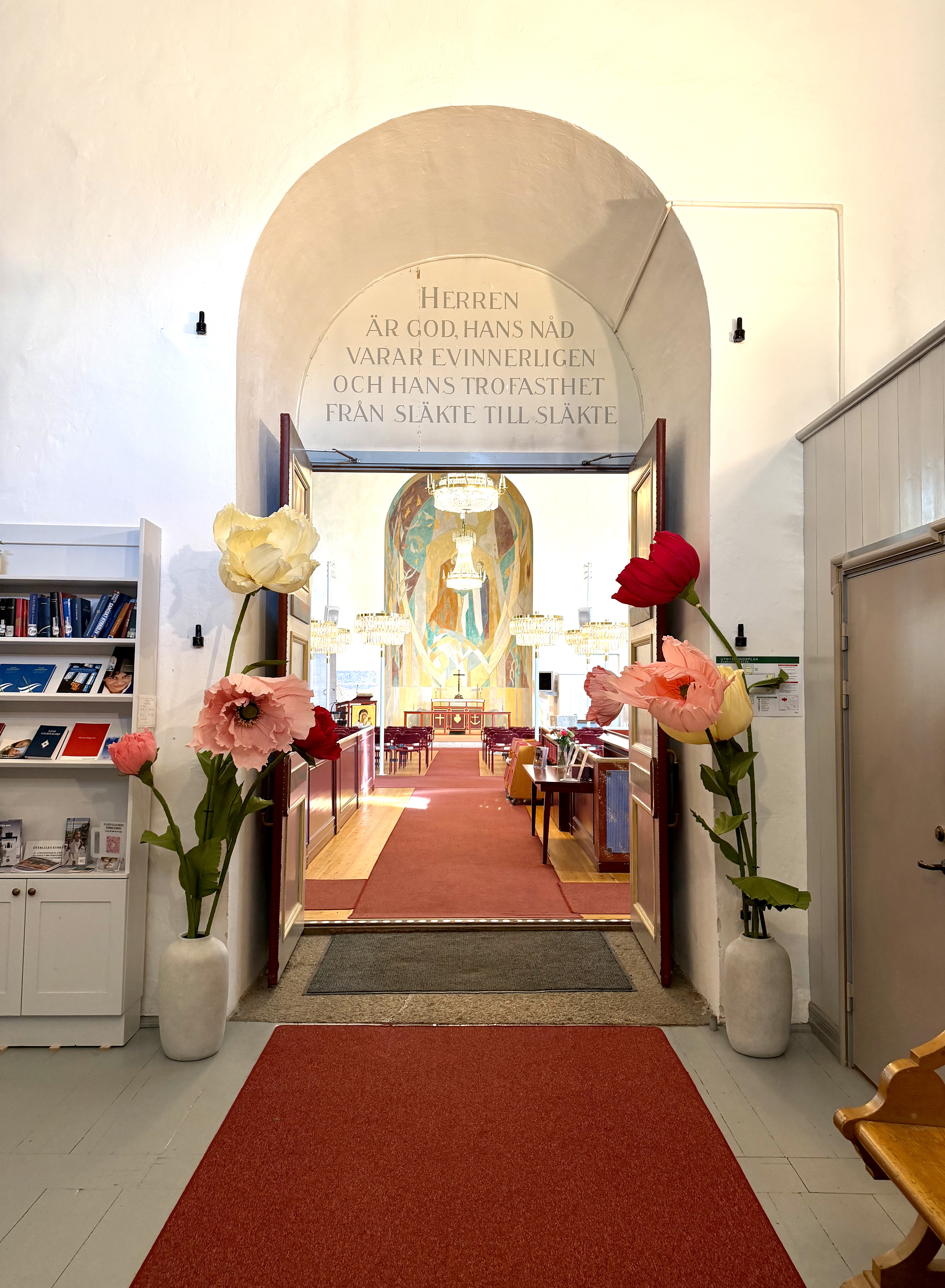 Vy från entrén in mot kyrkorummet i Överluleå kyrka. På båda sidor om dörröppningen står stora dekorativa blommor i vita vaser. Genom de öppna dörrarna syns röda kyrkbänkar, kristallkronor och koret med ett färgstarkt konstverk. Ovanför dörren finns en inskription med bibelord.
