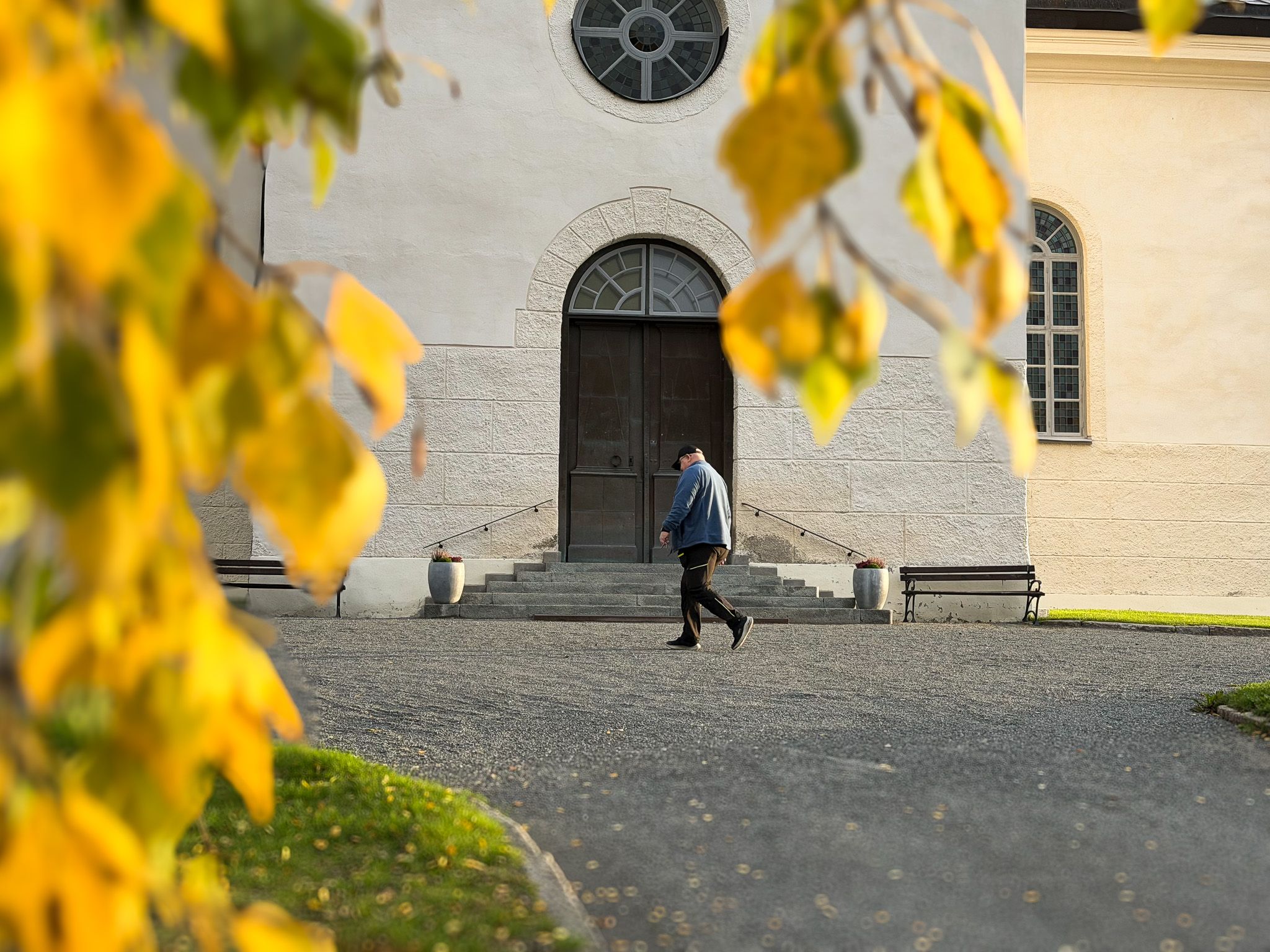 Höstlöv i gula nyanser ramar in en kyrkobyggnad. En person går över den öppna gårdsplanen framför kyrkans entré med trappa, bänkar och stora dörrar. Soligt dagsljus och höstfärger präglar bilden.