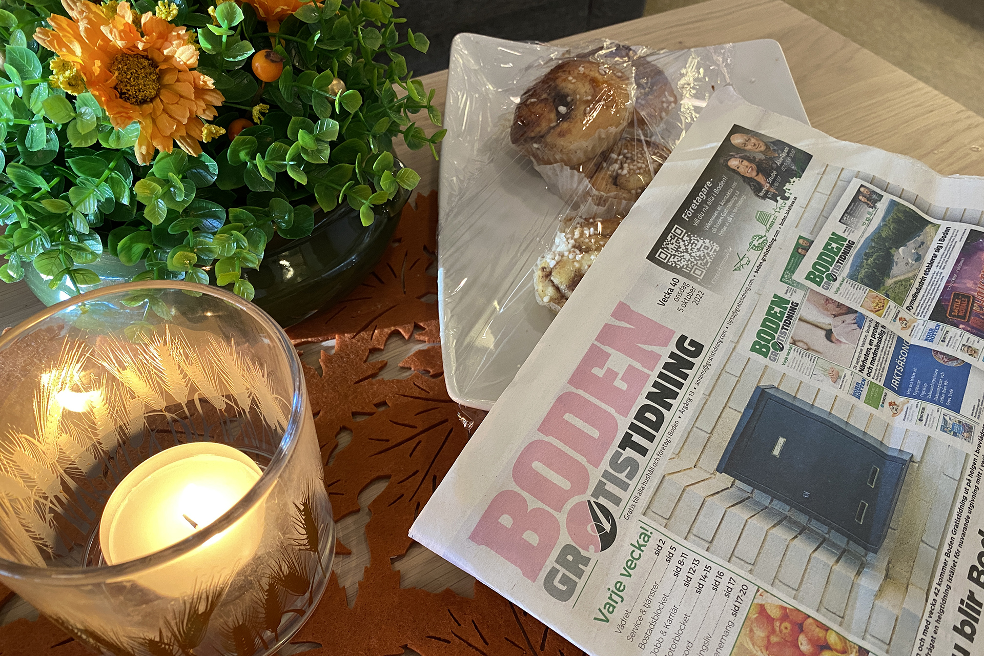 Ett bord med levande ljus, blommor, tidning och ett fat med bullar.
