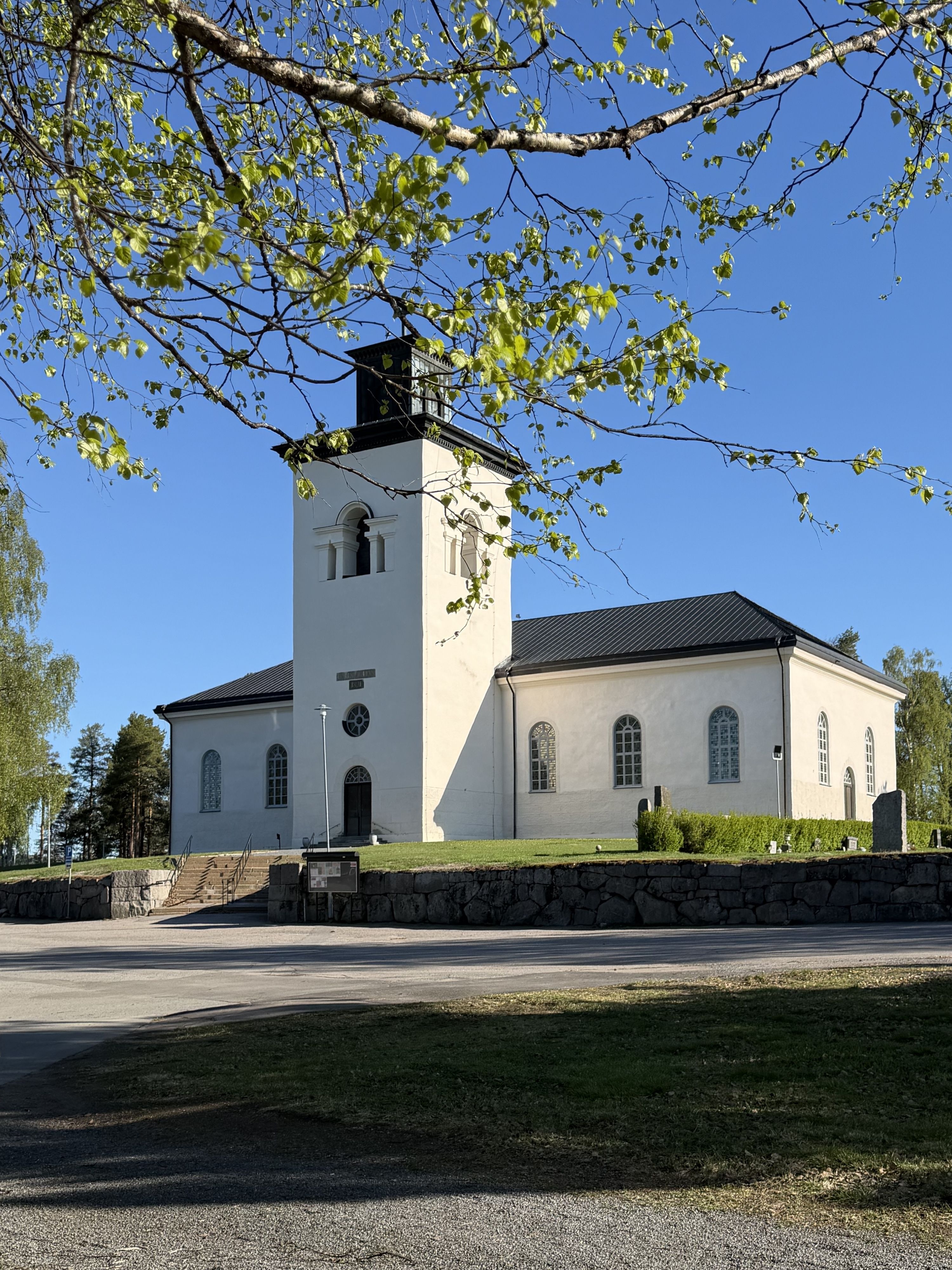 Överluleå kyrka i vårsol. närmast i bild ser man björkgrenar med nyutslagna blad. I bakgrunden en vit kyrka med svart tak.