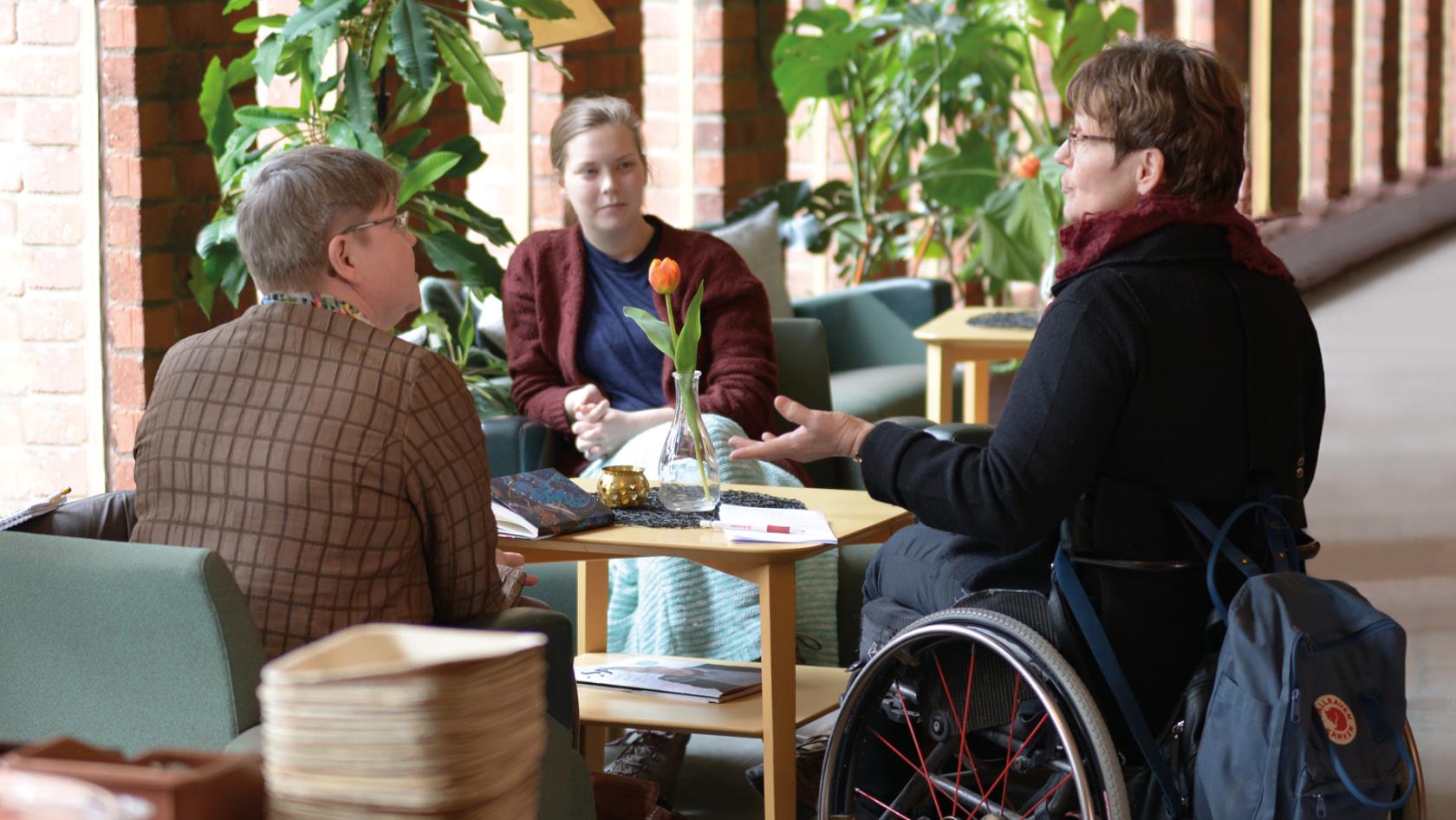 Tre personer sitter och pratar runt ett bord. En av dem sitter i rullstol. 