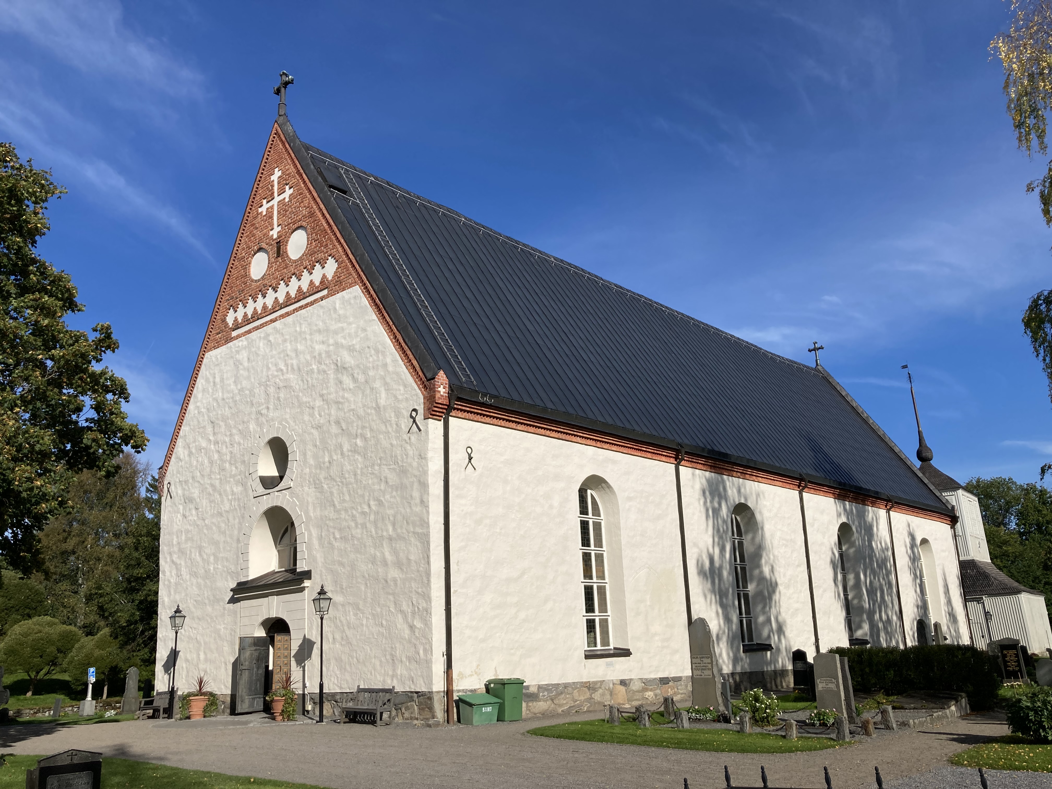 En vit medeltida kyrka. Enskeppig. Gavelspetsarna i rött tegel. Svart plåttak. Himlen är blå, och träden omkring börjar skifta lite i till höstfärger. 