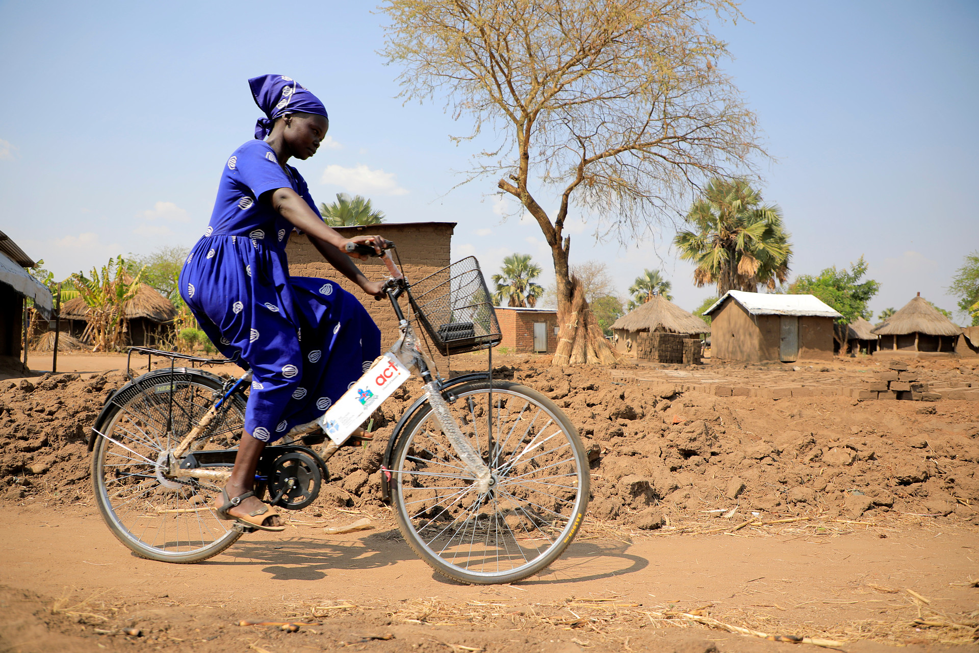 En kvinna i blå klänning cyklar på en grusväg i Afrika. På cykeln finns en röd-vit Act-skylt.
