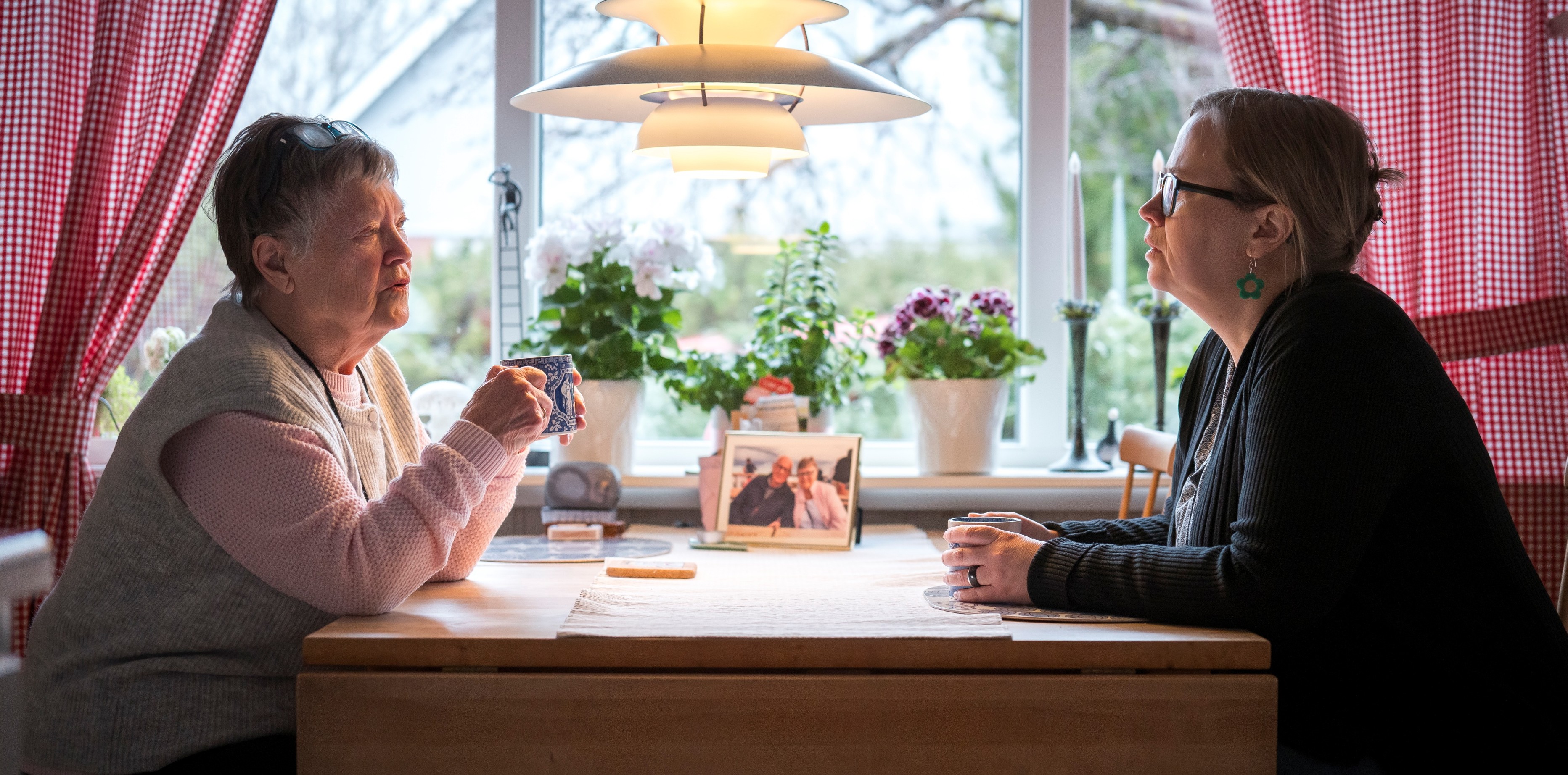 Två kvinnor sitter vid ett köksbord och dricker kaffe. Rödrutiga gardiner i fönstret. Trevlig atmosfär.