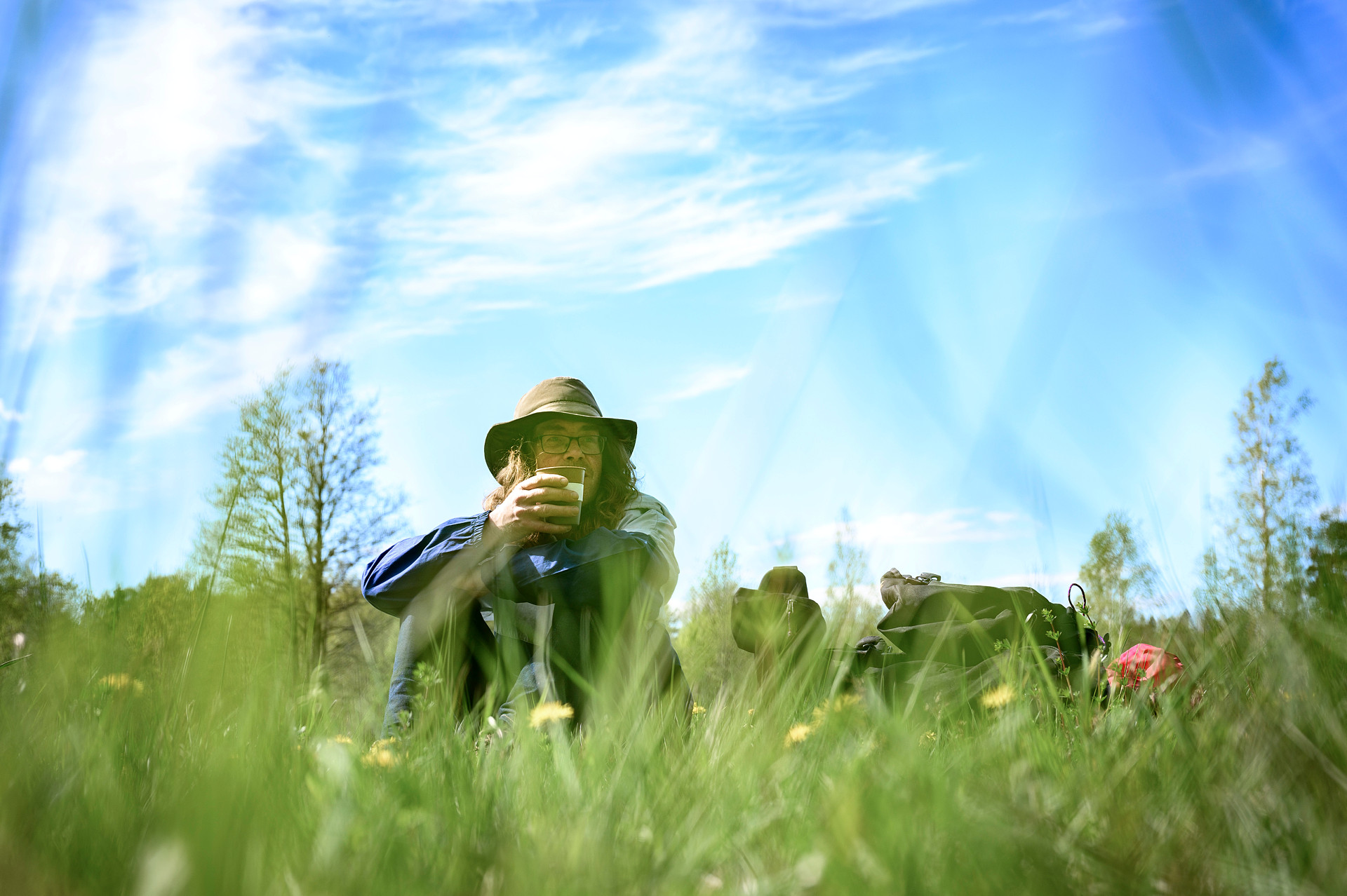 En man i hatt sitter i gräset och dricker kaffe. Himmlen är blå och nan ser ut att njuta. 