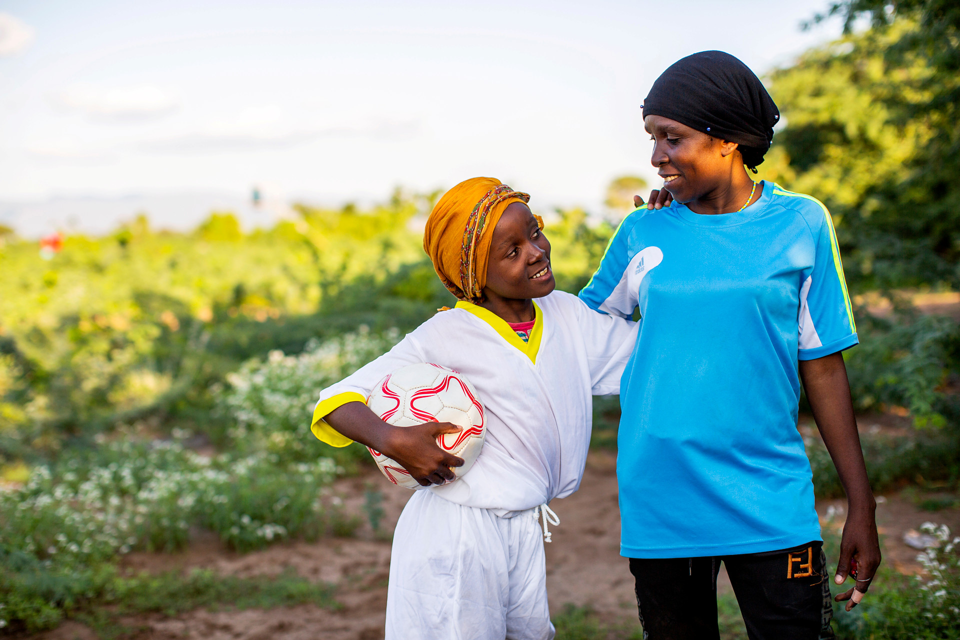 Bild på en kvinna i ljusblå tröja som tittar i ögonen på en flicka som står till vänster om henne på bilden. Flickan har ett vitt matchställ på sig och håller i en fotboll under ena armen. De båda håller om varandra med en varsin arm. 
