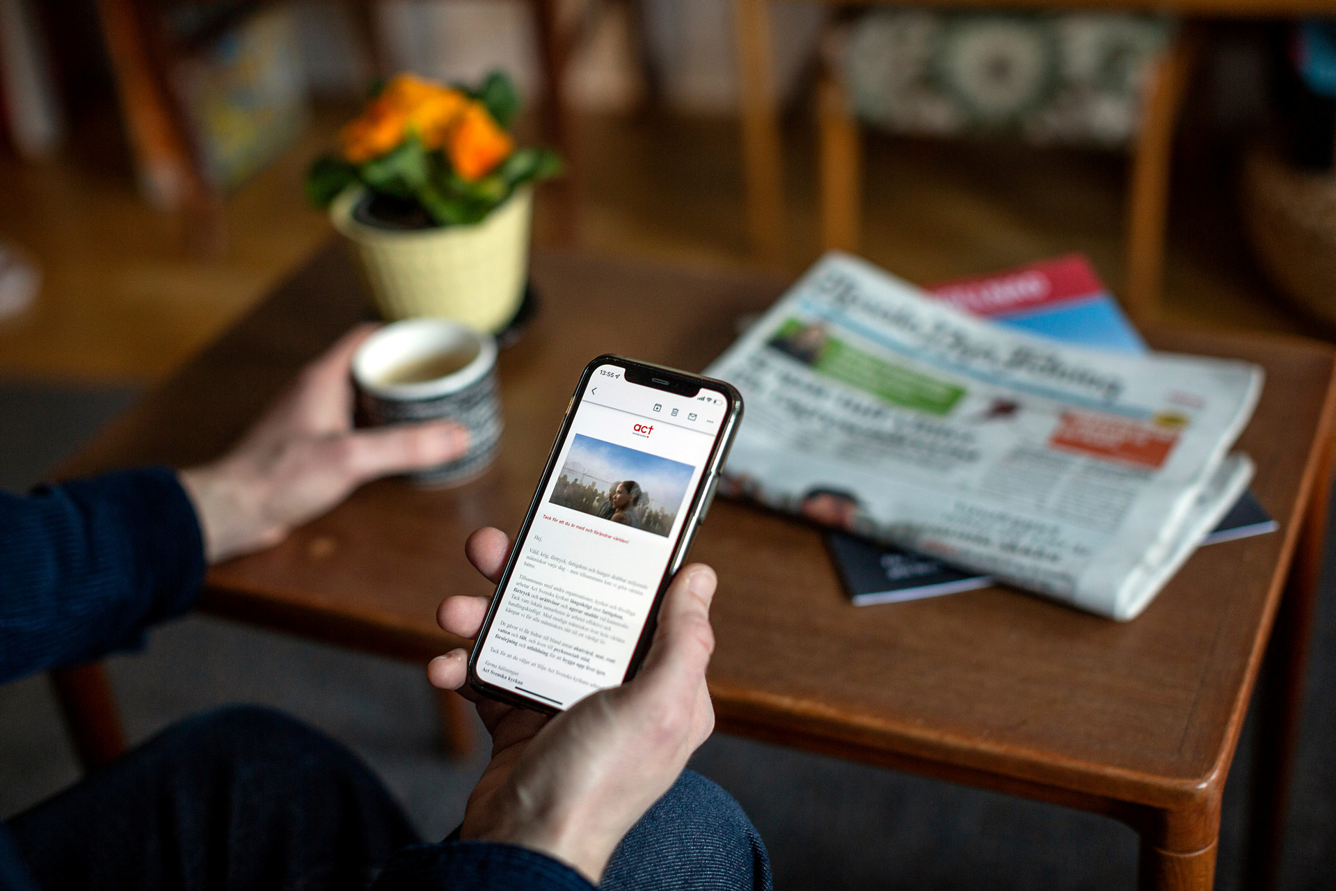 Ser en hand som håller i en mobiltelefon och läser ett nyhetsbrev. På soffbordet bakom står en kaffekopp och en liten hög med papperstidningar. 