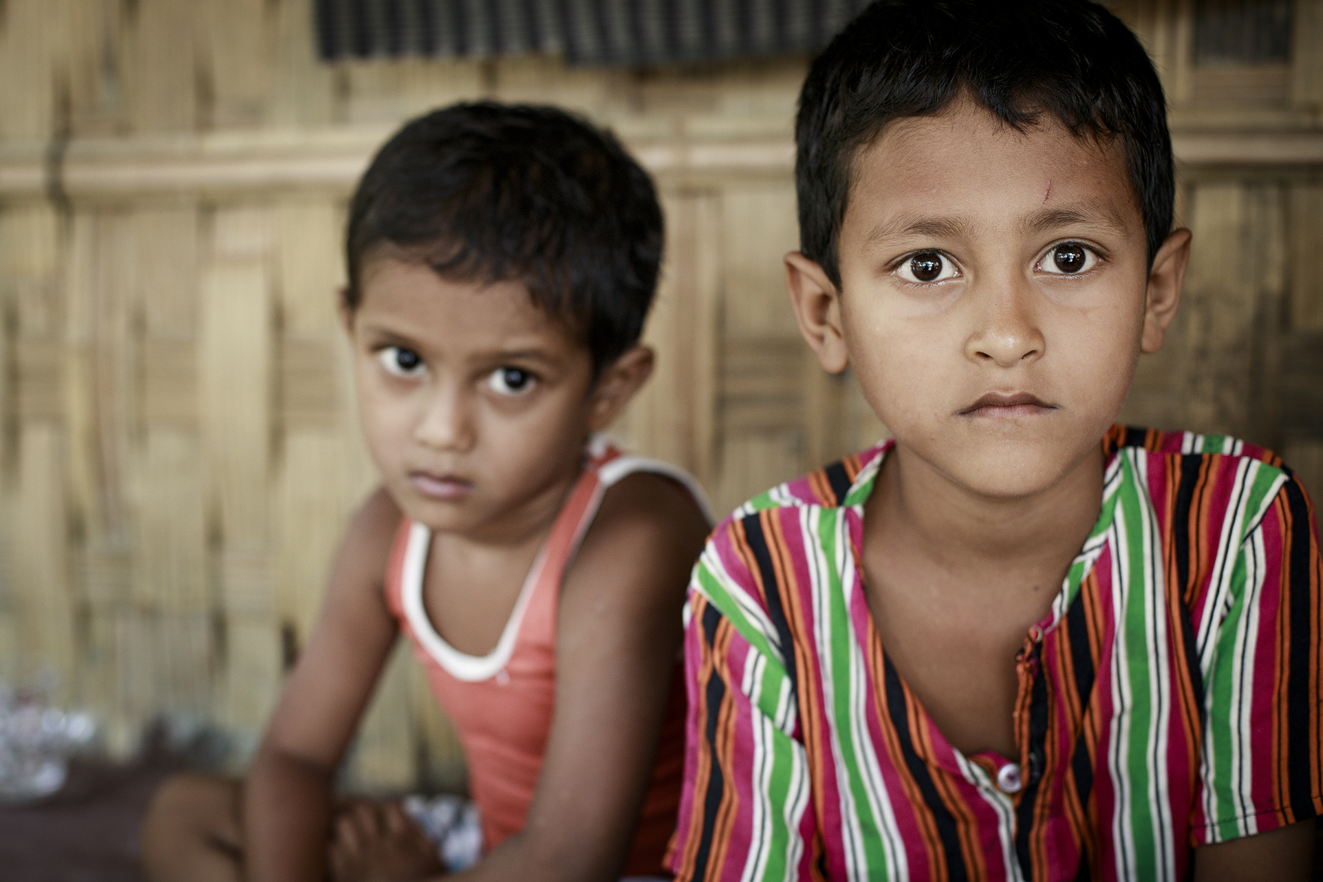 Två små pojkar tittar på något precis bakom kameran. Pojken längst fram är i fokus i bilden, han har en randig skjorta på sig. Bojken bakom har ett rött linne. De ser lite bekymmrade ut.  