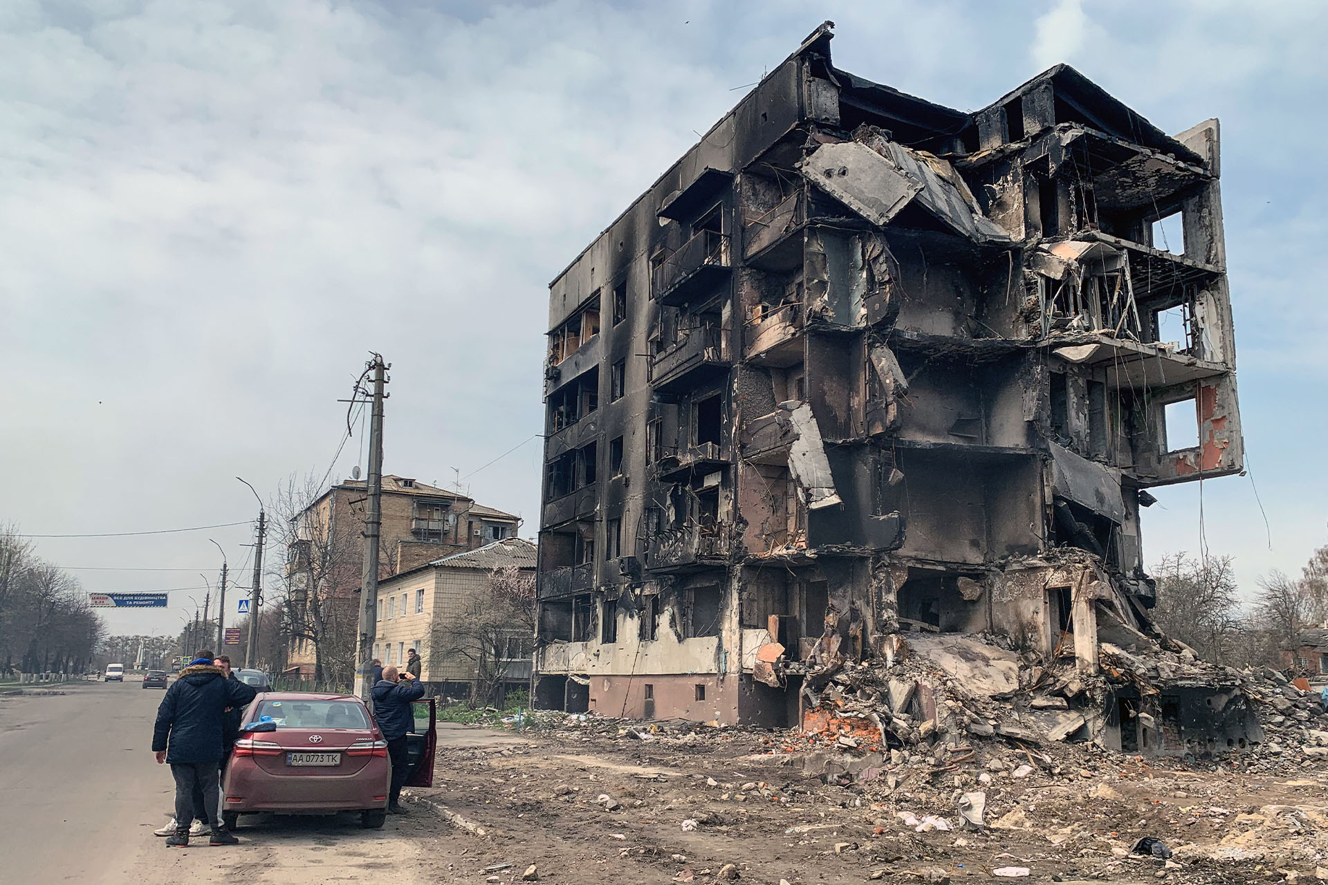 Bild på ett hus som förstörts i bombningarna. I förgrunden står ett antal personer vid en parkerad bil och tittar på det trasiga huset.