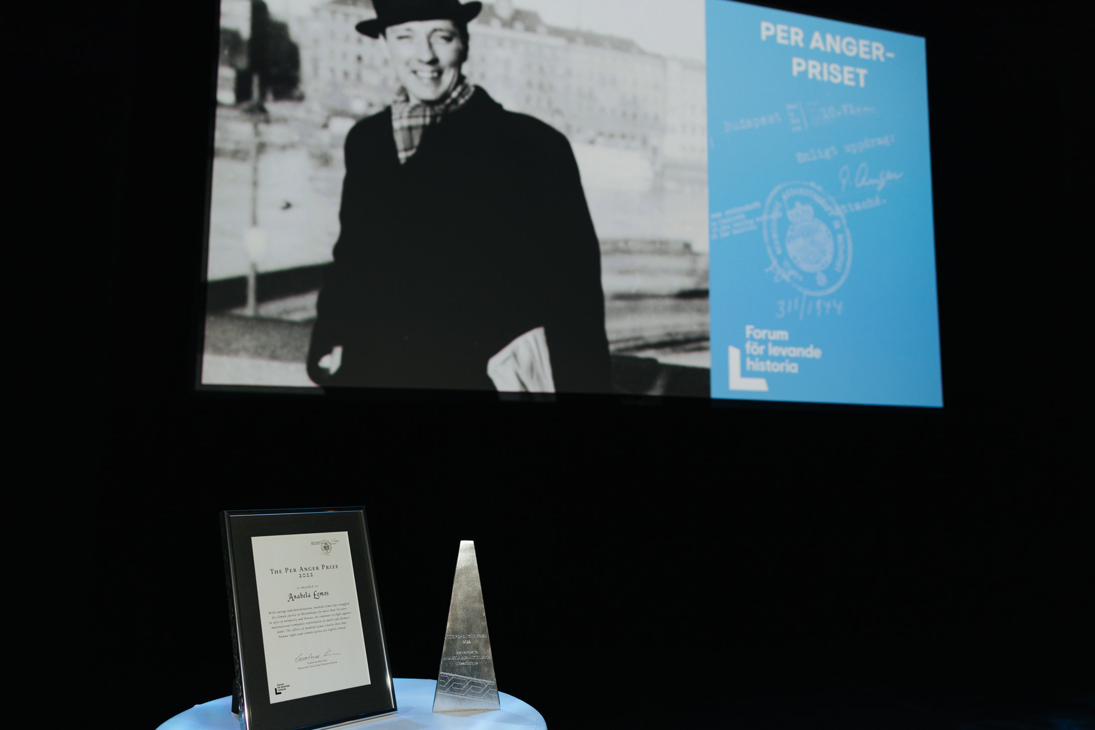 På ett bord står ett diplom och ett pris, på en skärm visas en bild av Per Anger