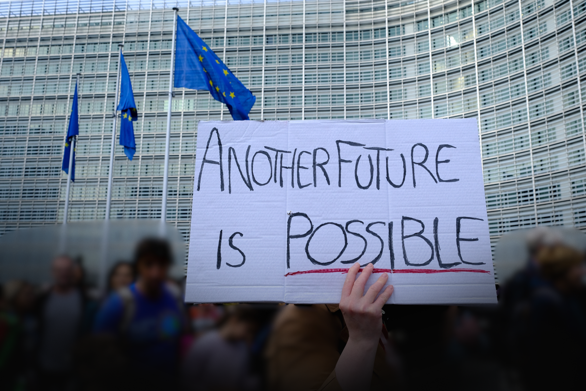 Folkmassa framför myndighetsbyggnad och EU-flaggor. I förgrunden ett kartongplakat med text Another future is possible