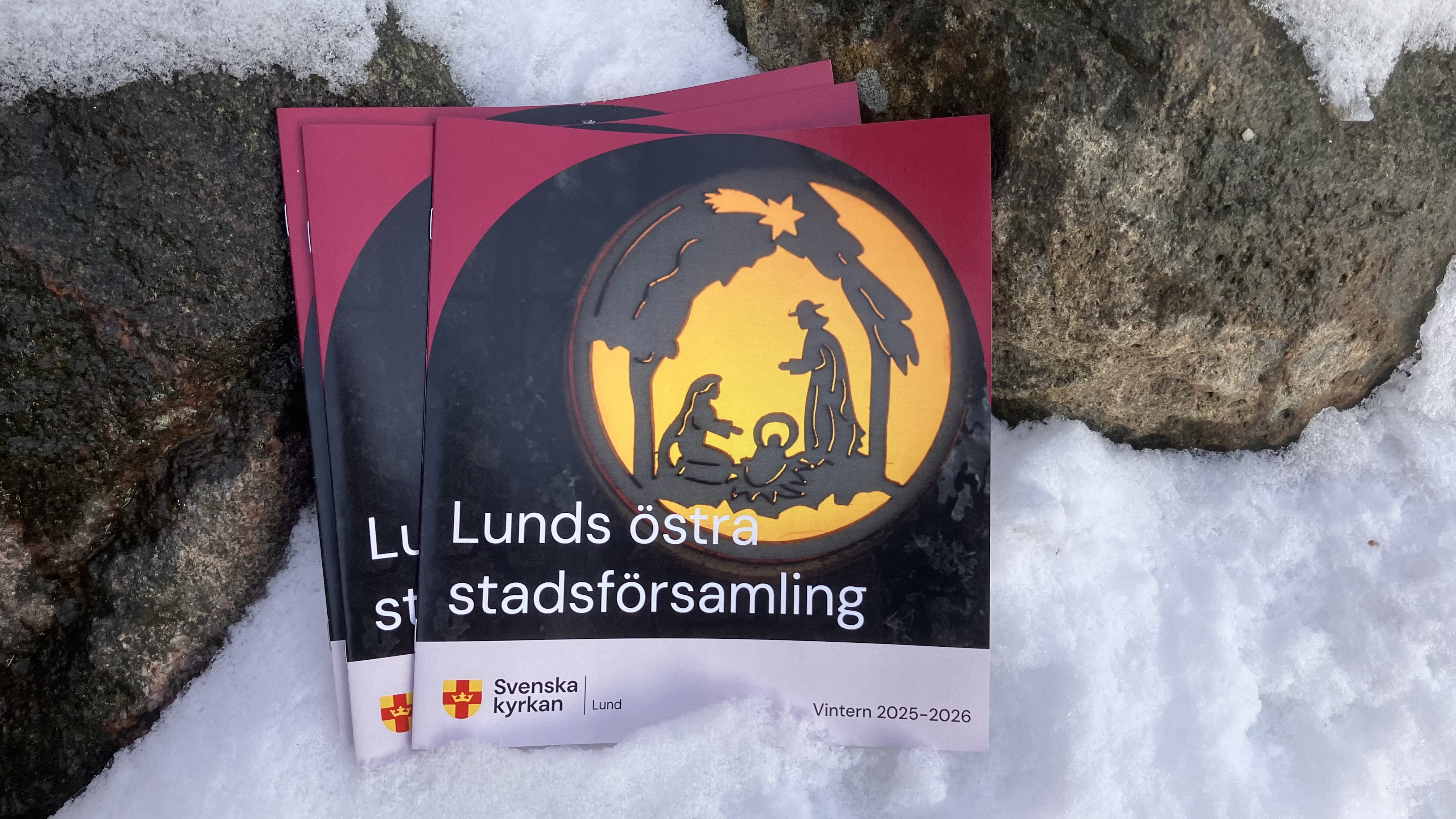 Några n4 2025 församlingsblad från Lunds östra stadsförsamling står lutade mot en sten med snö på