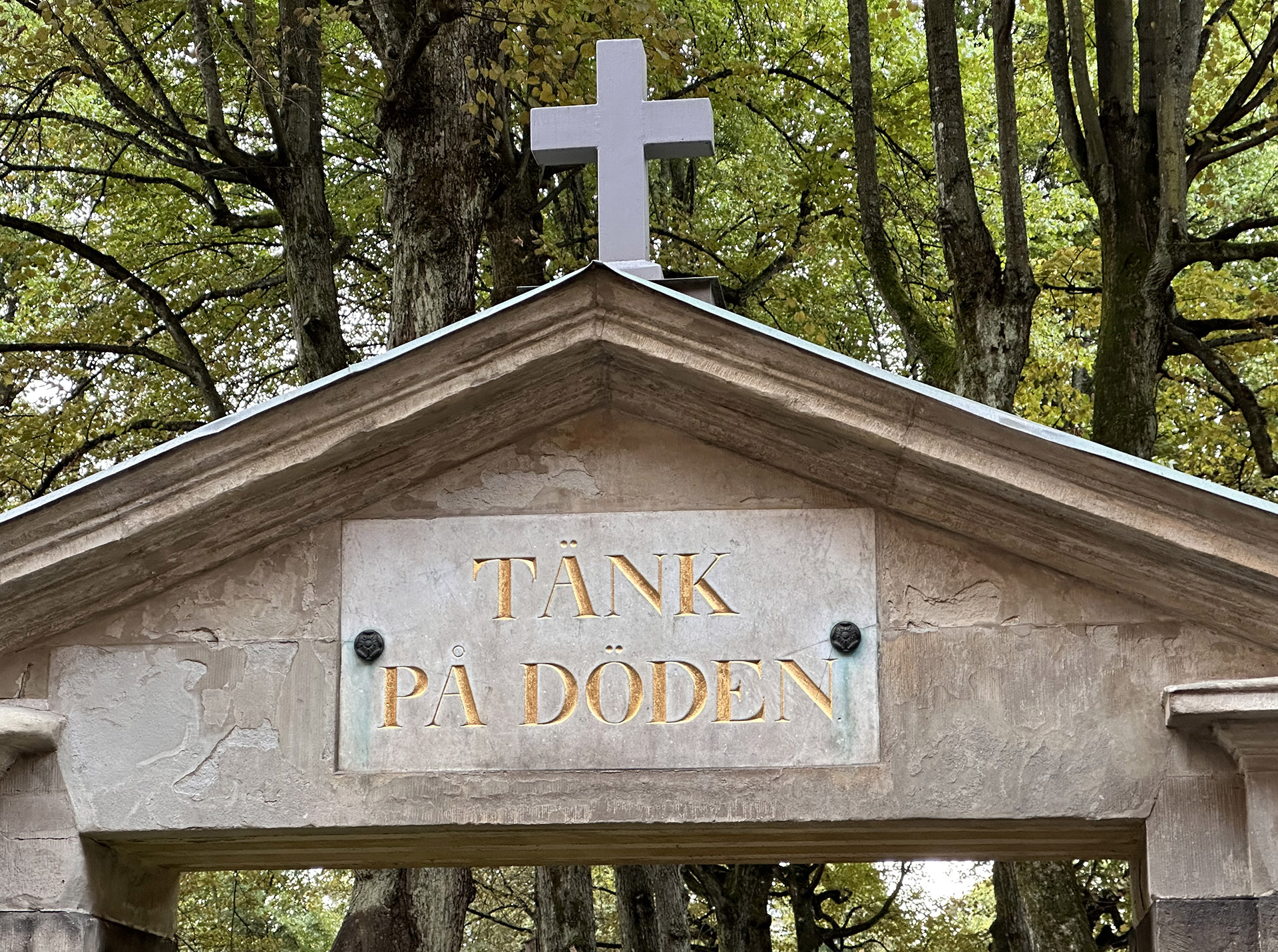 Stenportal med ett kors på toppen och en platta med inskriptionen Tänk på döden. I bakgrunden stora lövträd.