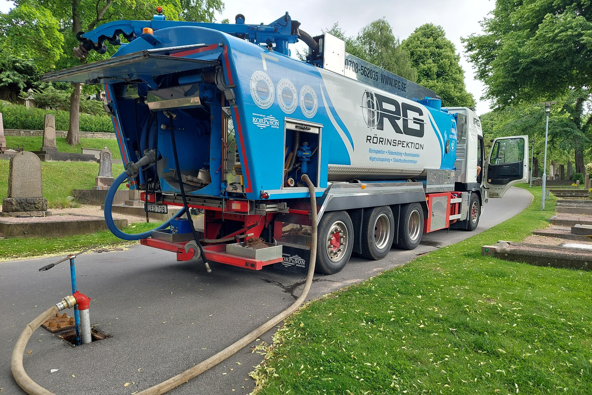 Spolbil parkerad på gata på kyrkgård, slang i förgrunden och träd och gravar i bakgrunden.