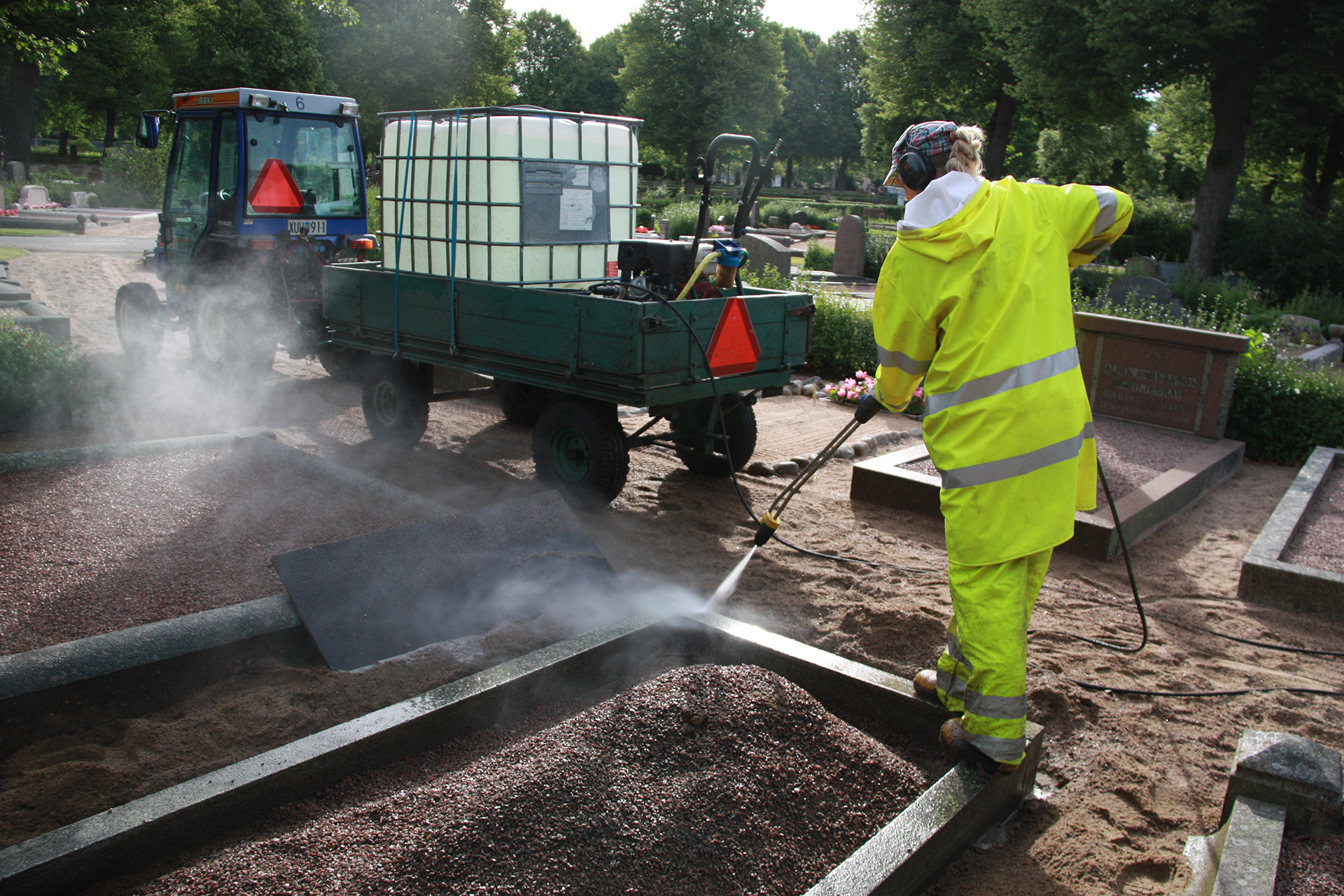 Person i neongula regnkläder tvättar en gravram med en högtrycksspruta på en kyrkogård.