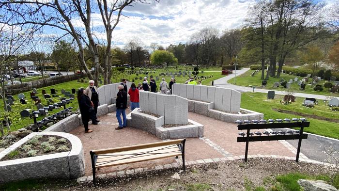 Flertal personer står vid bänk, ljushållare och granitpelare på grusad del av kyrkogård.