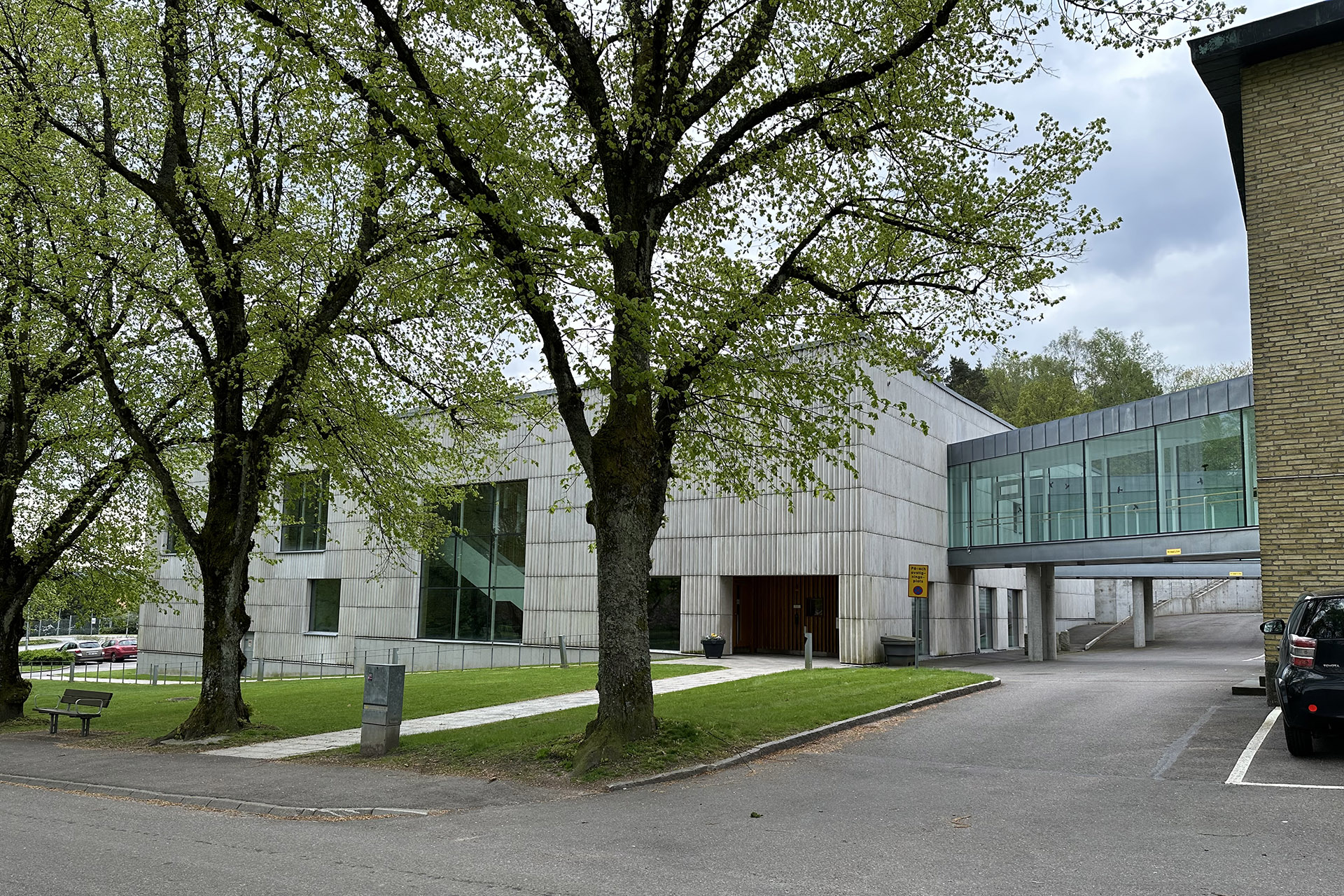 Grå byggnad med en inglasad övergång till en gul tegelbyggnad. I förgrunden grönskande träd och en asfalterad väg.