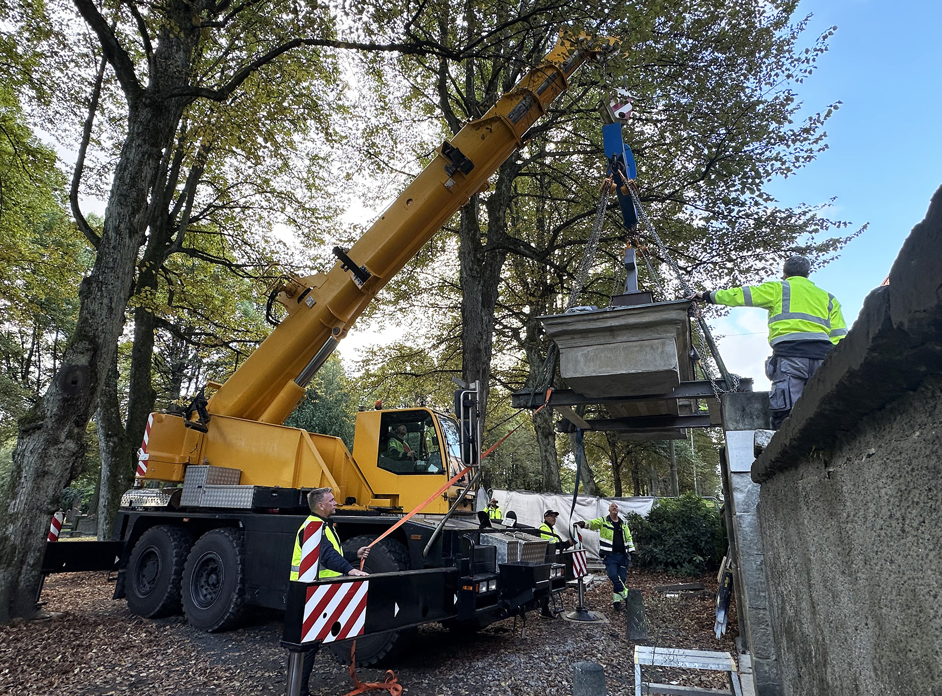 Lyftkran lyfter stenportal på plats uppe på pelare på kyrkogård med träd runtomkring.
