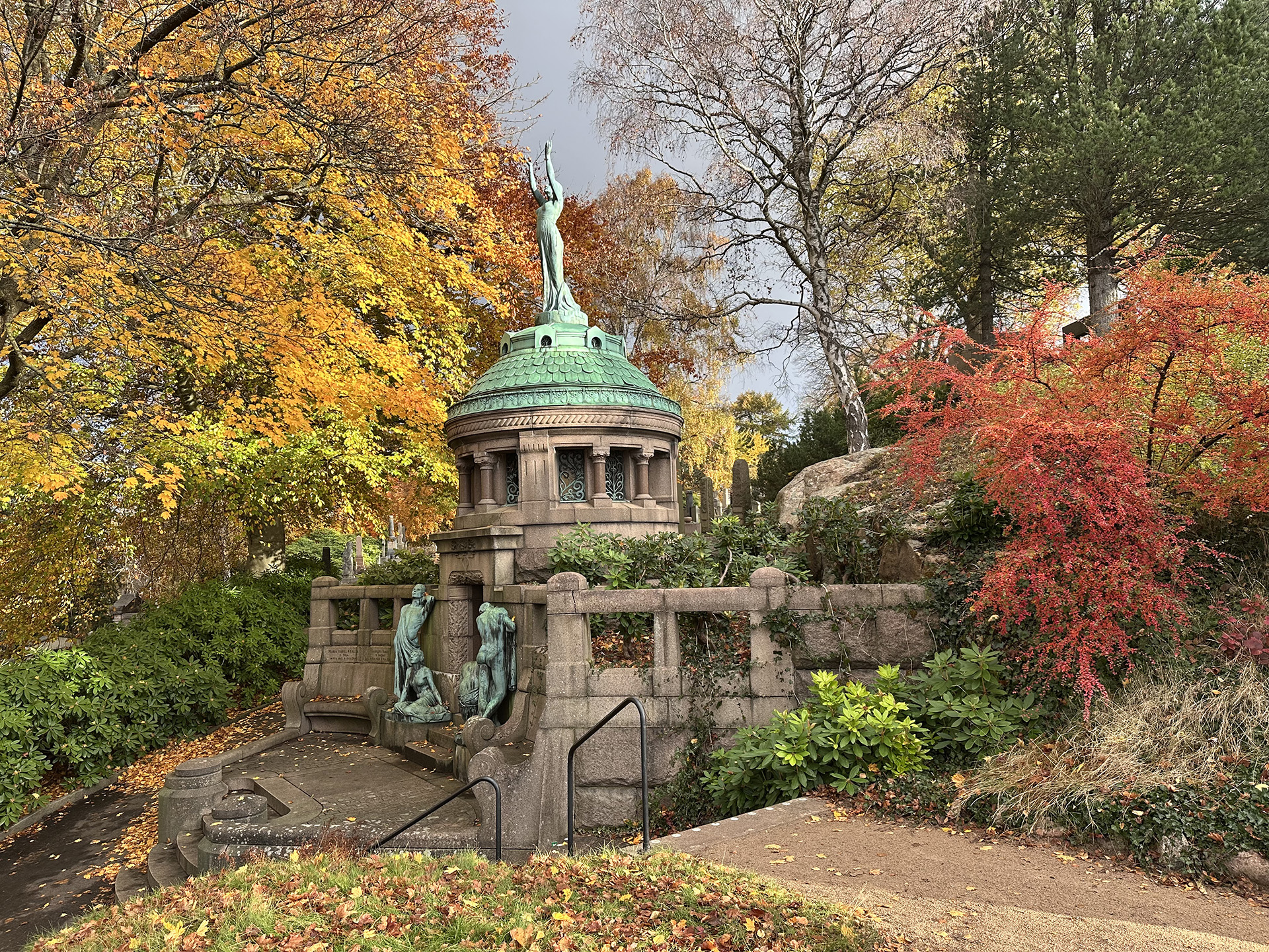 Stort gravmonument i sten med skulptur på toppen omgiven av träd med löv i höstens alla färger.
