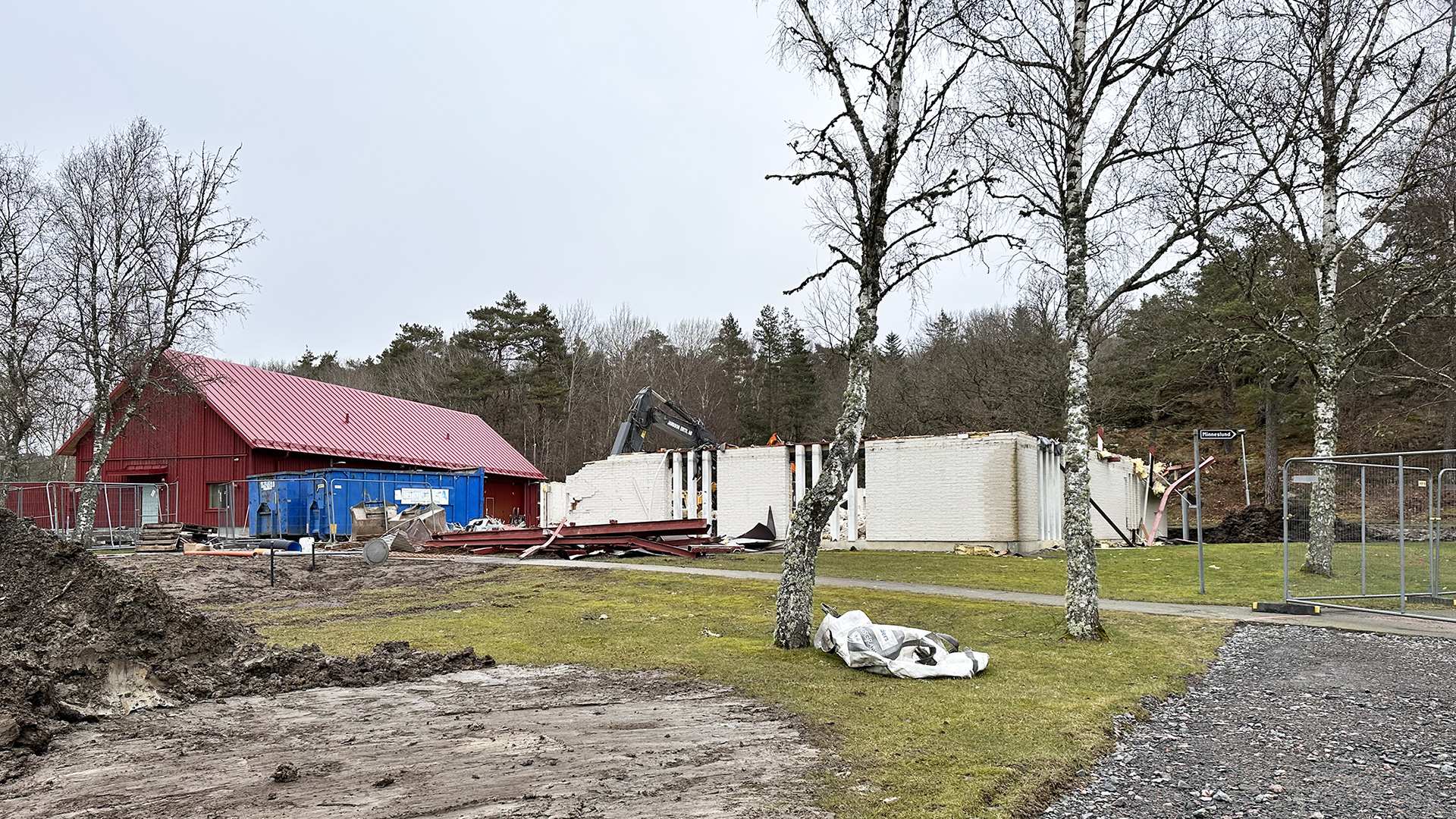 Halvt riven vit tegelbyggnad med träd i förgrunden och en röd ekonomibyggnad i bakgrunden.