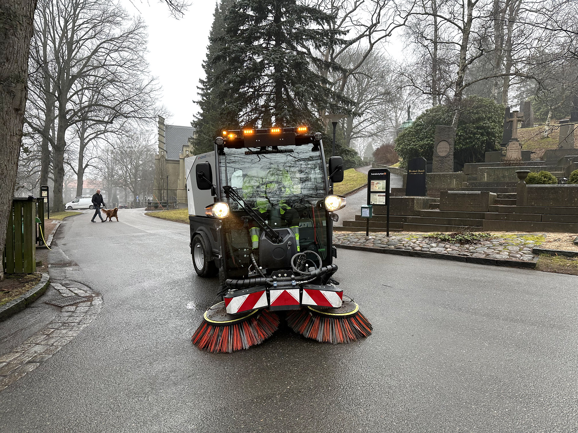 Sopmaskin kör mot fotografen på asfalterad väg på begravningsplats. I bakgrunden träd, en person med hund och ett kapell.