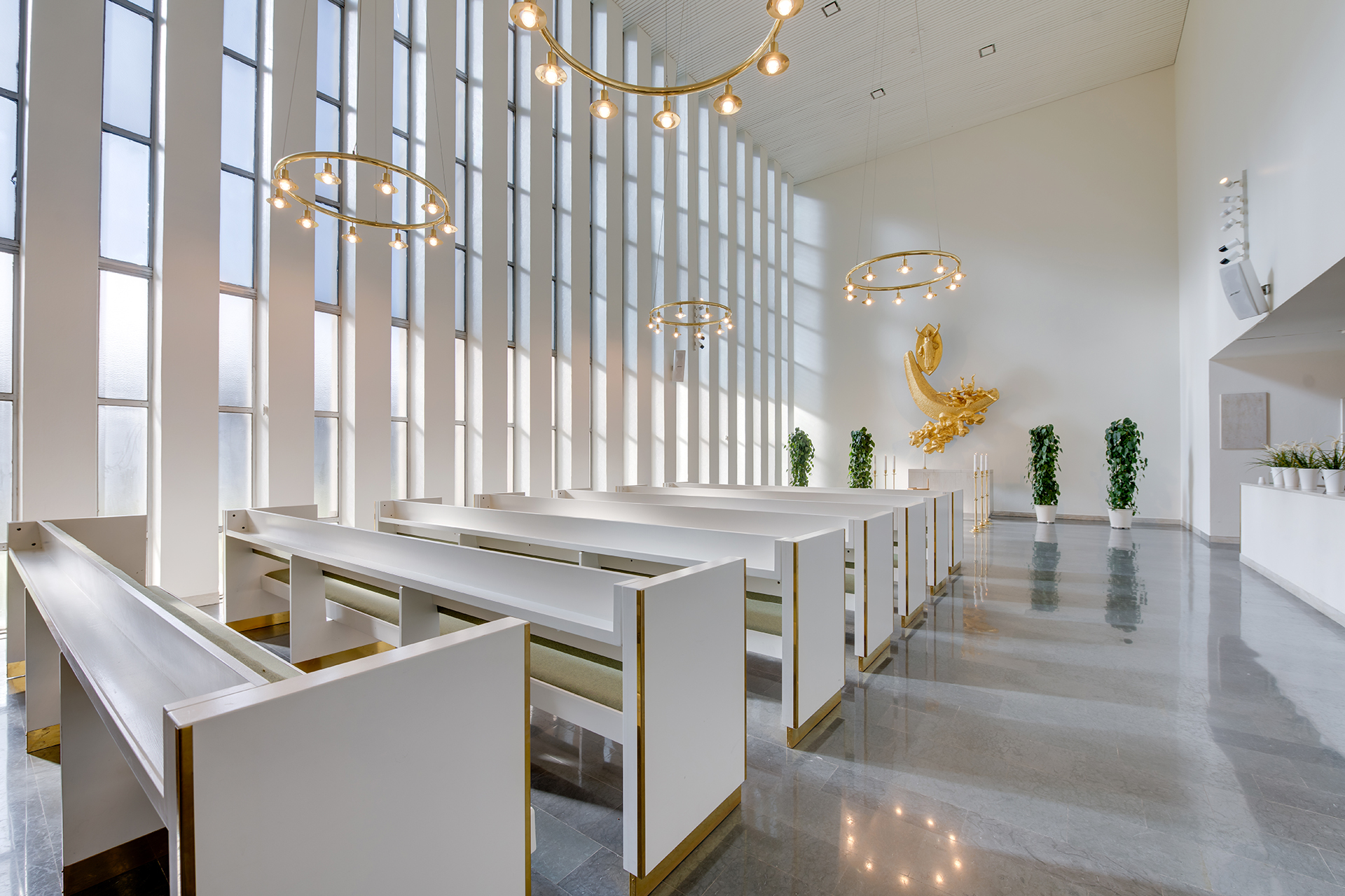 Bänkrader i lokal med högt i tak och stora fönster. I fonden en förgylld väggskulptur.