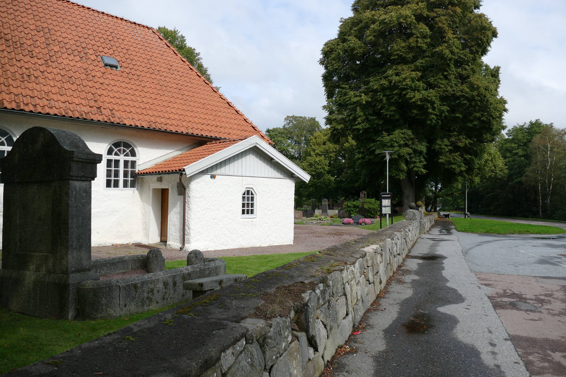 kyrkobyggnad med stenmur framför och grönskande träd i bakgrunden.
