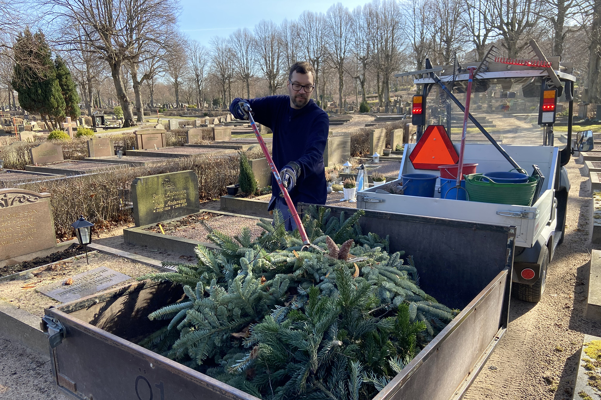 Man lyfter på gamla granrisprydnader på en släpkärra mellan gravarna på en kyrkogård.